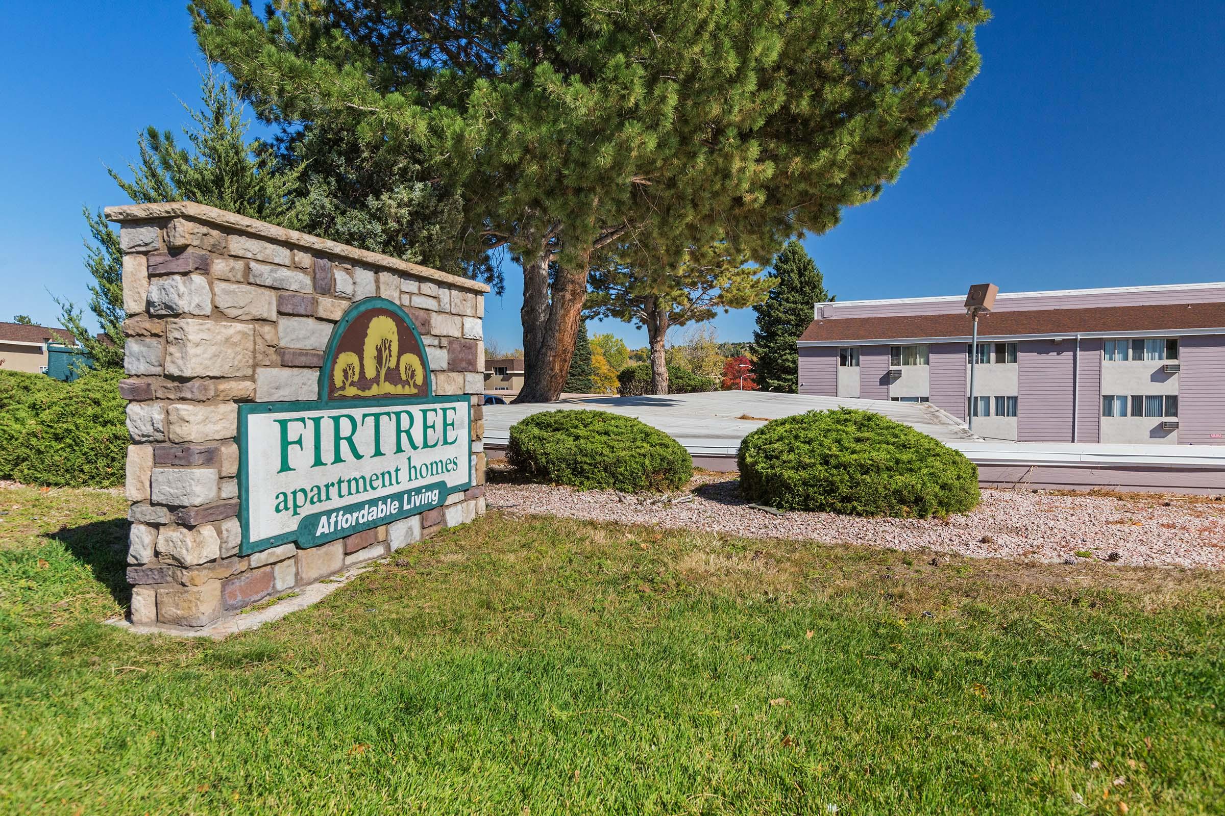 A stone sign for Fir Tree Apartment Homes, featuring the text "Fir Tree Apartment Homes" and "Affordable Living," surrounded by well-maintained grass and shrubs, with a multi-unit residential building visible in the background against a clear blue sky.