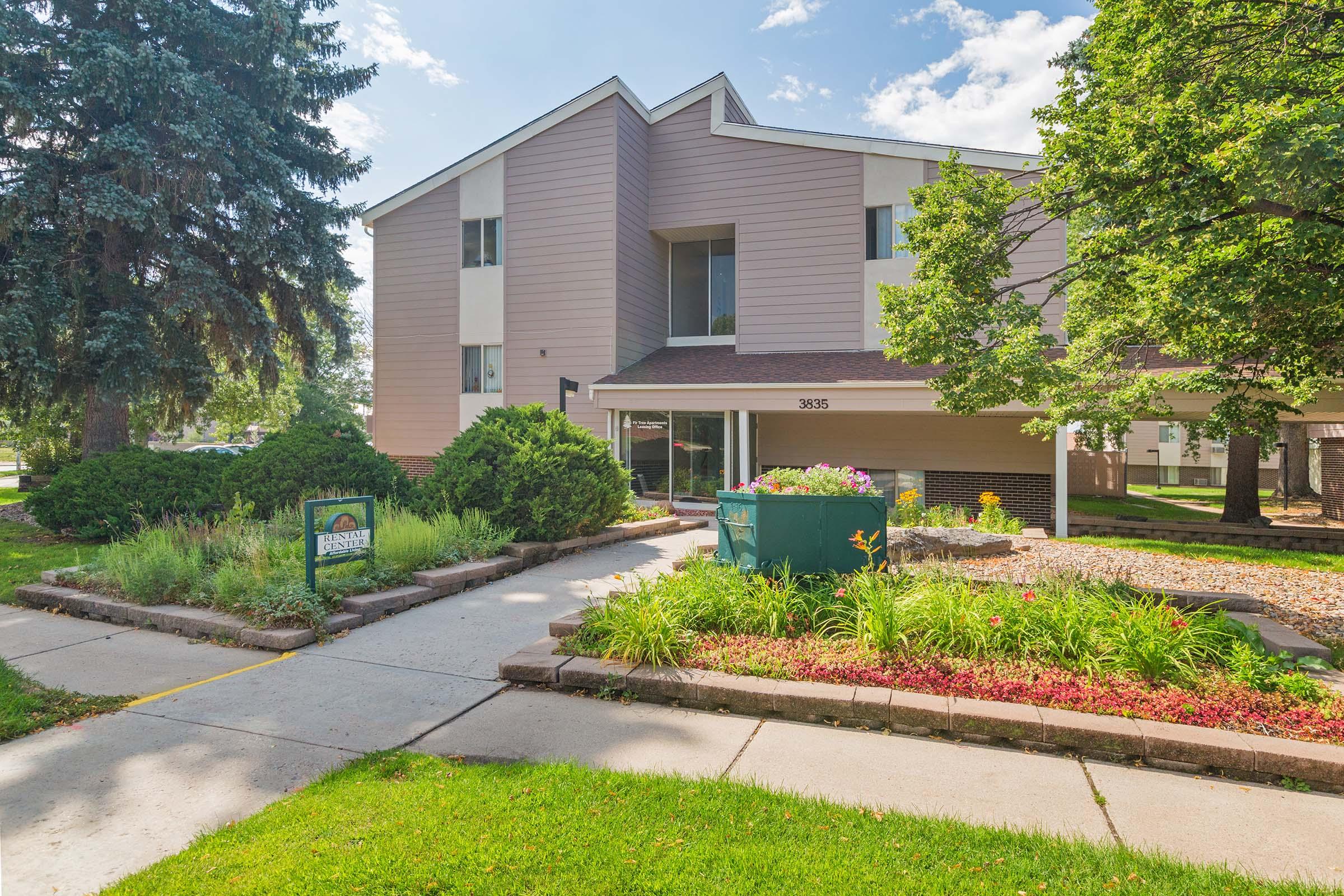 A well-maintained, modern apartment building with a light pink exterior. The entrance features a landscaped pathway lined with greenery and colorful flower beds. A sign near the walkway displays the building's address, and large trees provide shade to the surrounding area under a clear blue sky.