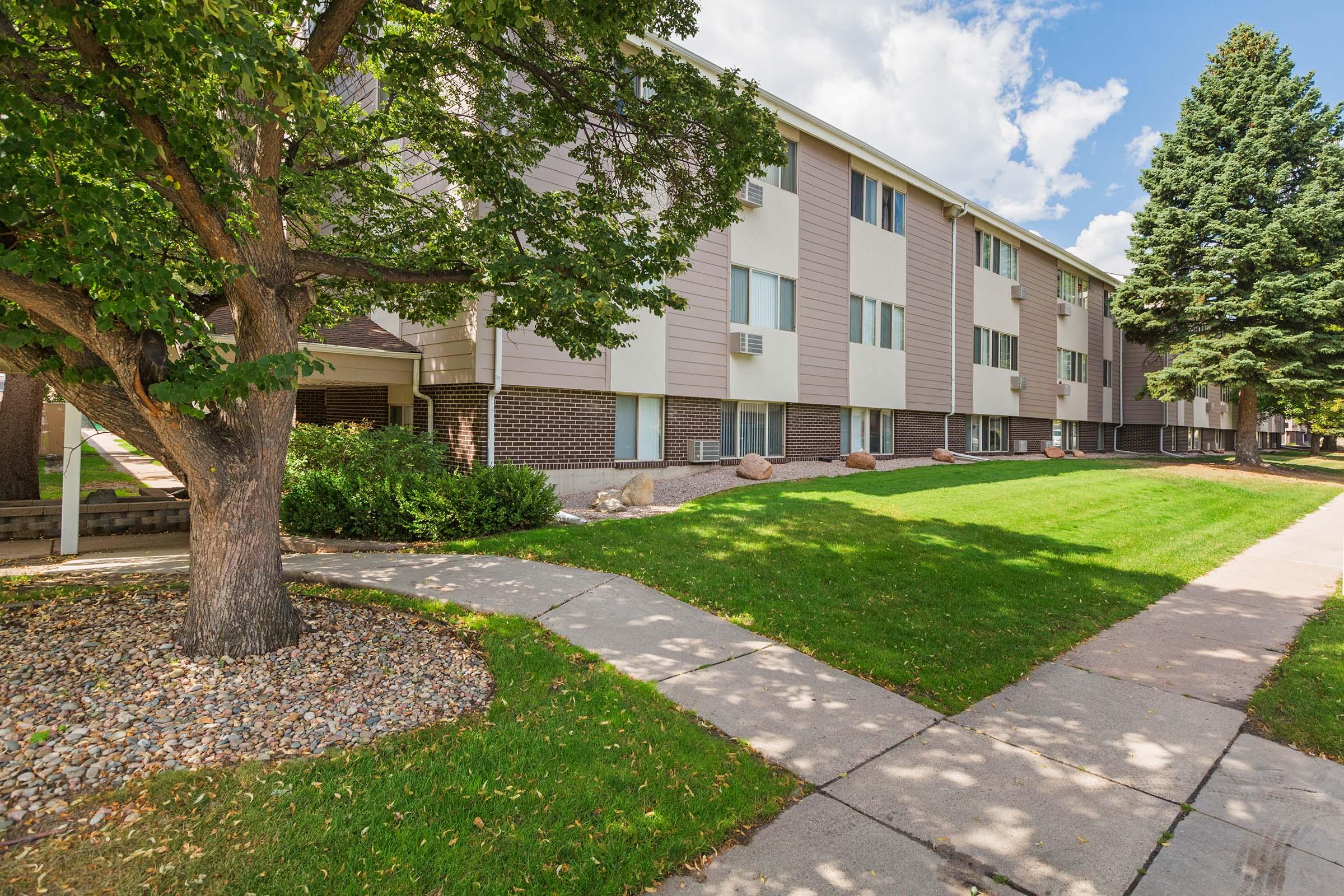 A well-maintained apartment building with multiple windows, set amidst a green lawn and a tree. The sidewalk leads up to the entrance, and there are decorative rocks along the edge of the lawn. The sky is partly cloudy, creating a pleasant outdoor atmosphere.