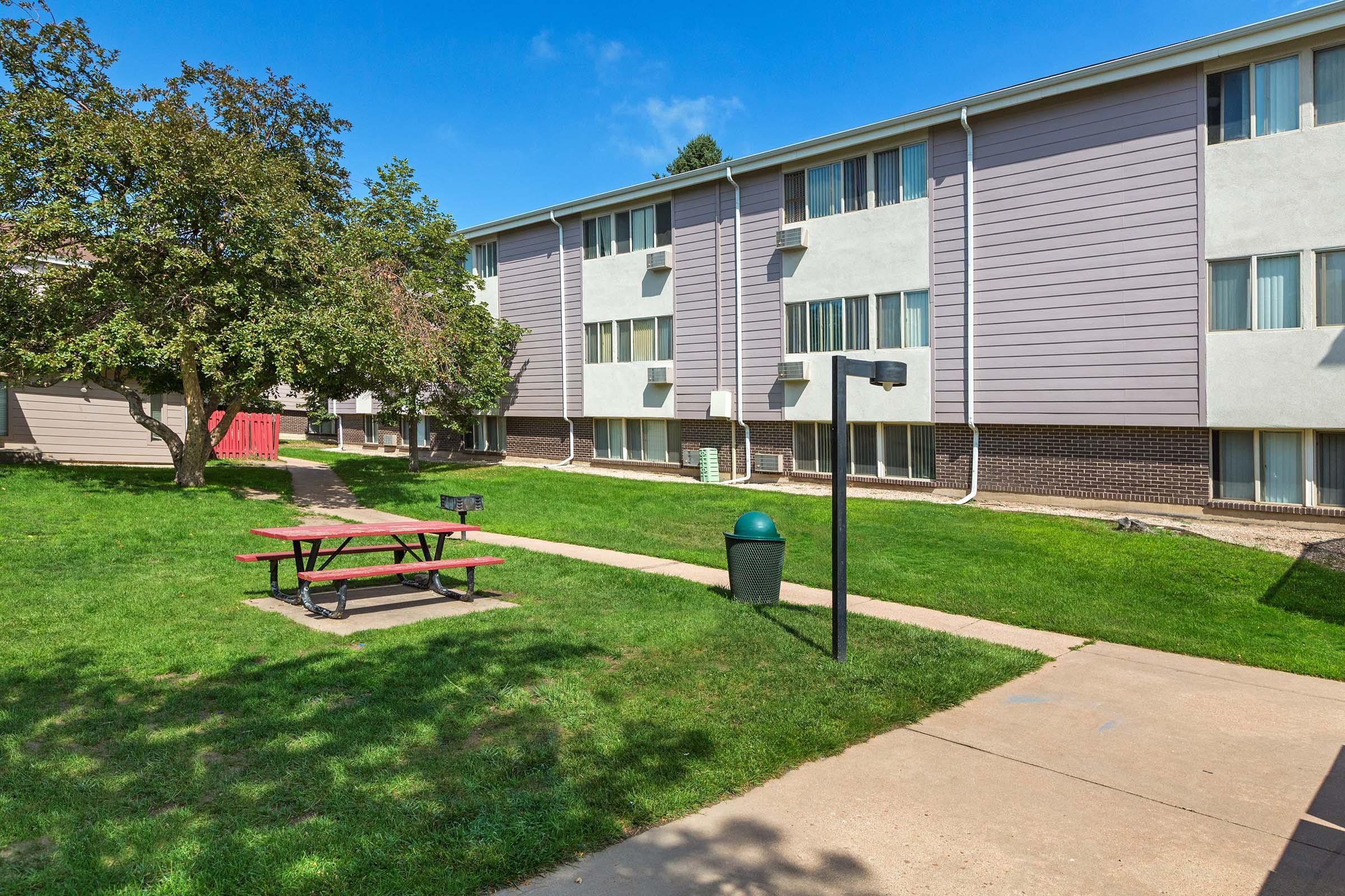 A landscaped area featuring a grassy courtyard with a picnic table and trash can, adjacent to a multi-story residential building. The building has a light exterior with large windows and is surrounded by trees under a clear blue sky.