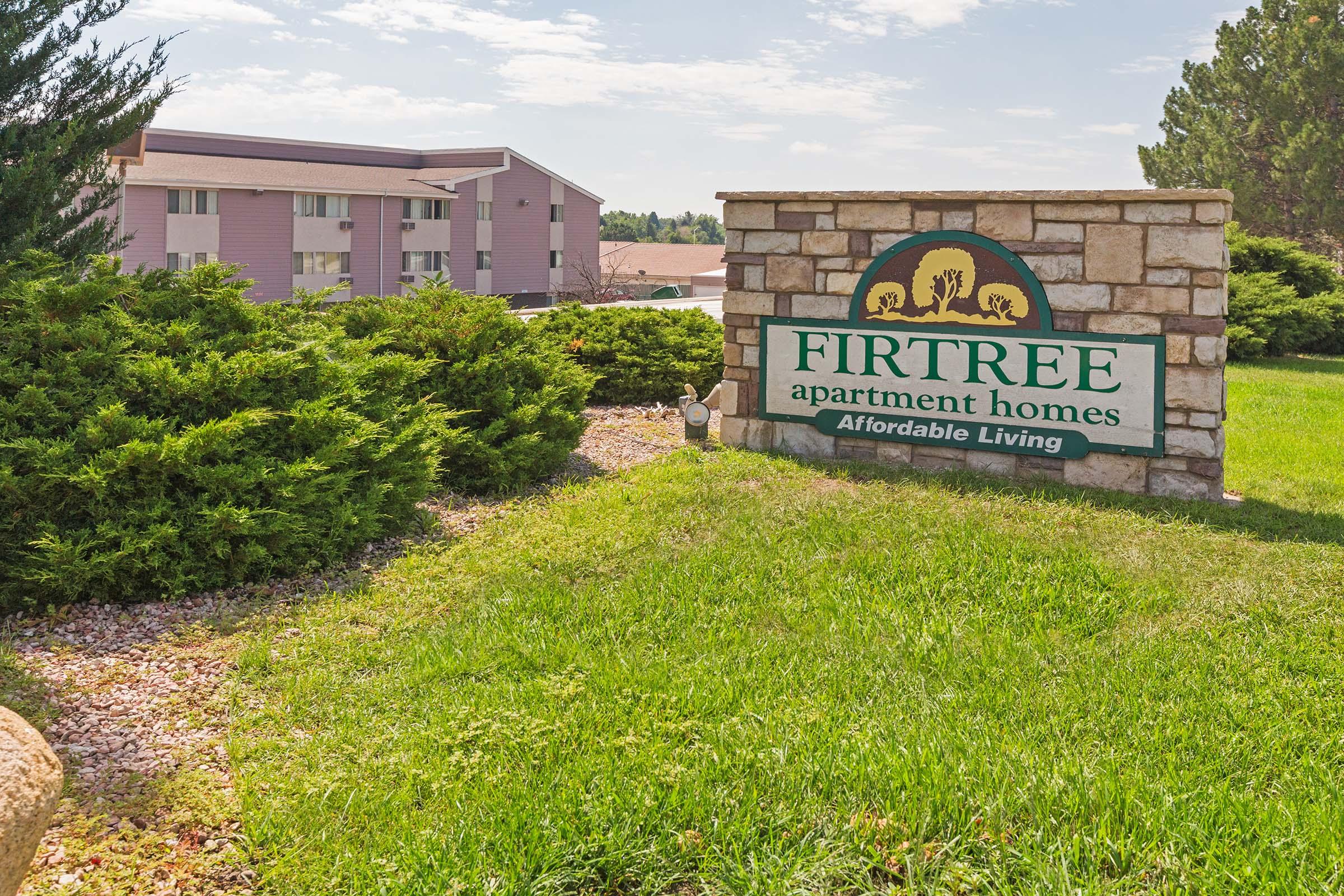 A stone sign for Fir Tree Apartment Homes, featuring a logo with trees and the text "Fir Tree Apartment Homes - Affordable Living." The sign is situated on a well-maintained green lawn with shrubs in the background. In the distance, there are pink apartment buildings under a partly cloudy sky.