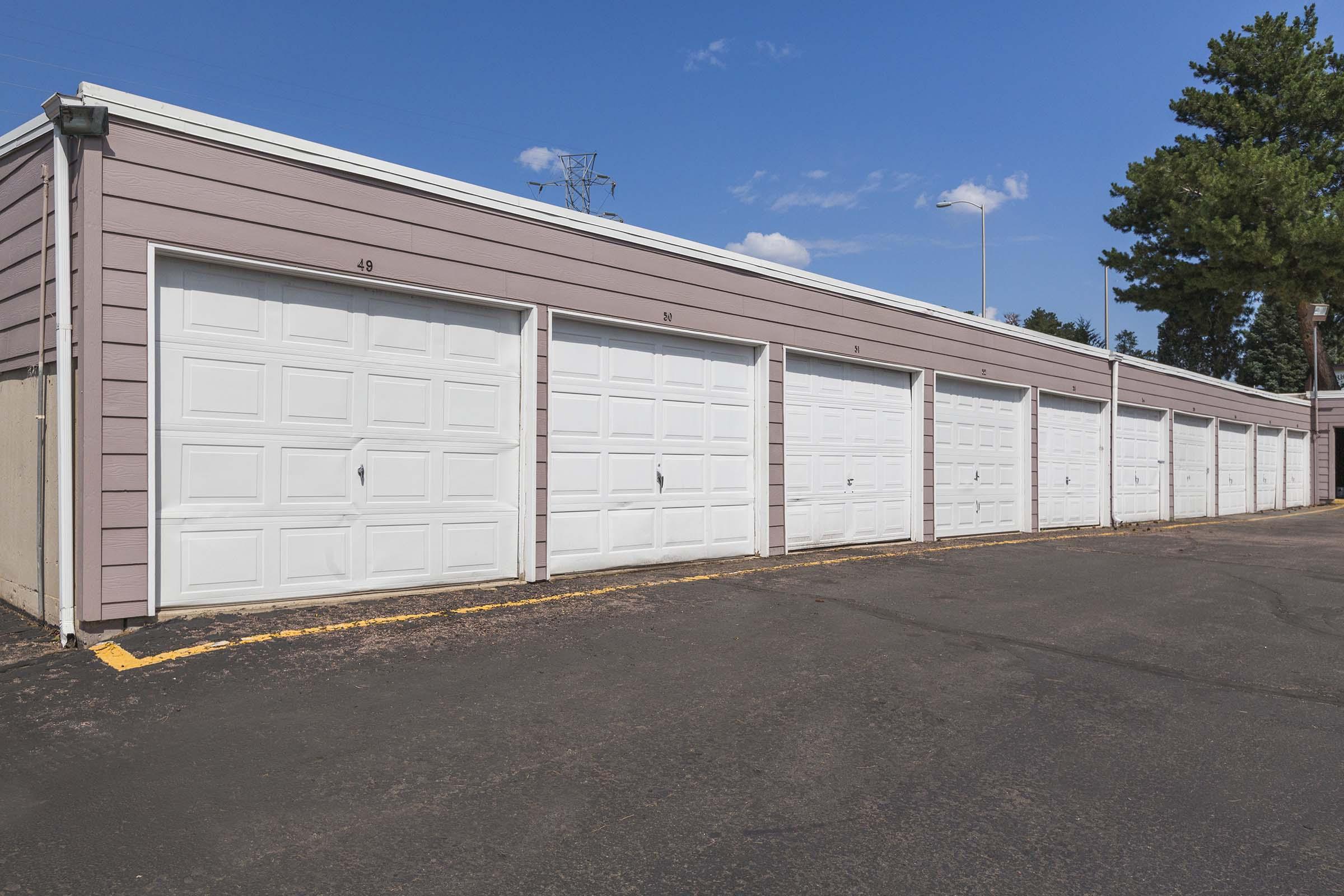 Row of white garage doors in a storage facility, with a clear blue sky in the background. The garages are numbered and situated on a paved parking area, surrounded by trees. Sunlight casts shadows, indicating a bright day.