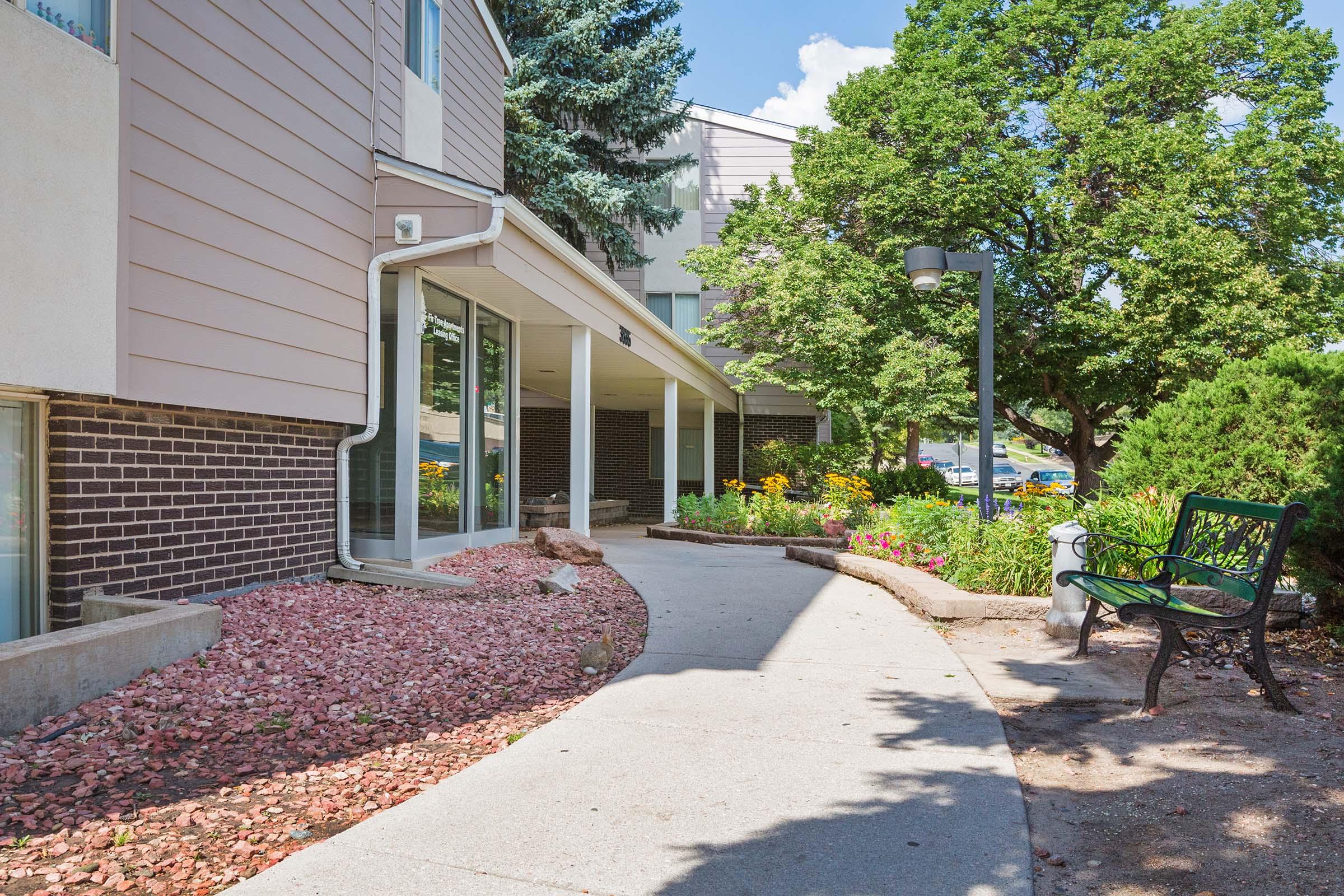 A walkway lined with flower beds leads to the entrance of a modern building. The path is surrounded by greenery, including trees and shrubs. A bench sits nearby, providing a place to sit and enjoy the outdoor space. The scene is bright and inviting, with a clear blue sky in the background.