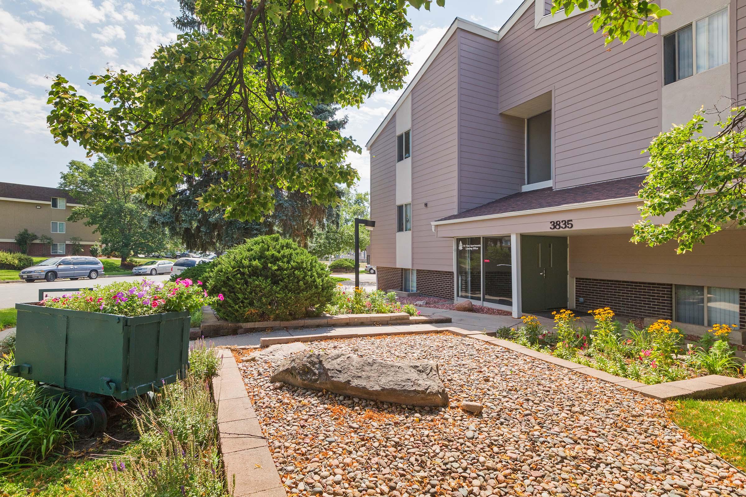 A landscaped outdoor area featuring a stone and gravel garden, colorful flowers, and a green utility cart. In the background, a modern building with light-colored siding and large windows is visible under a clear blue sky.