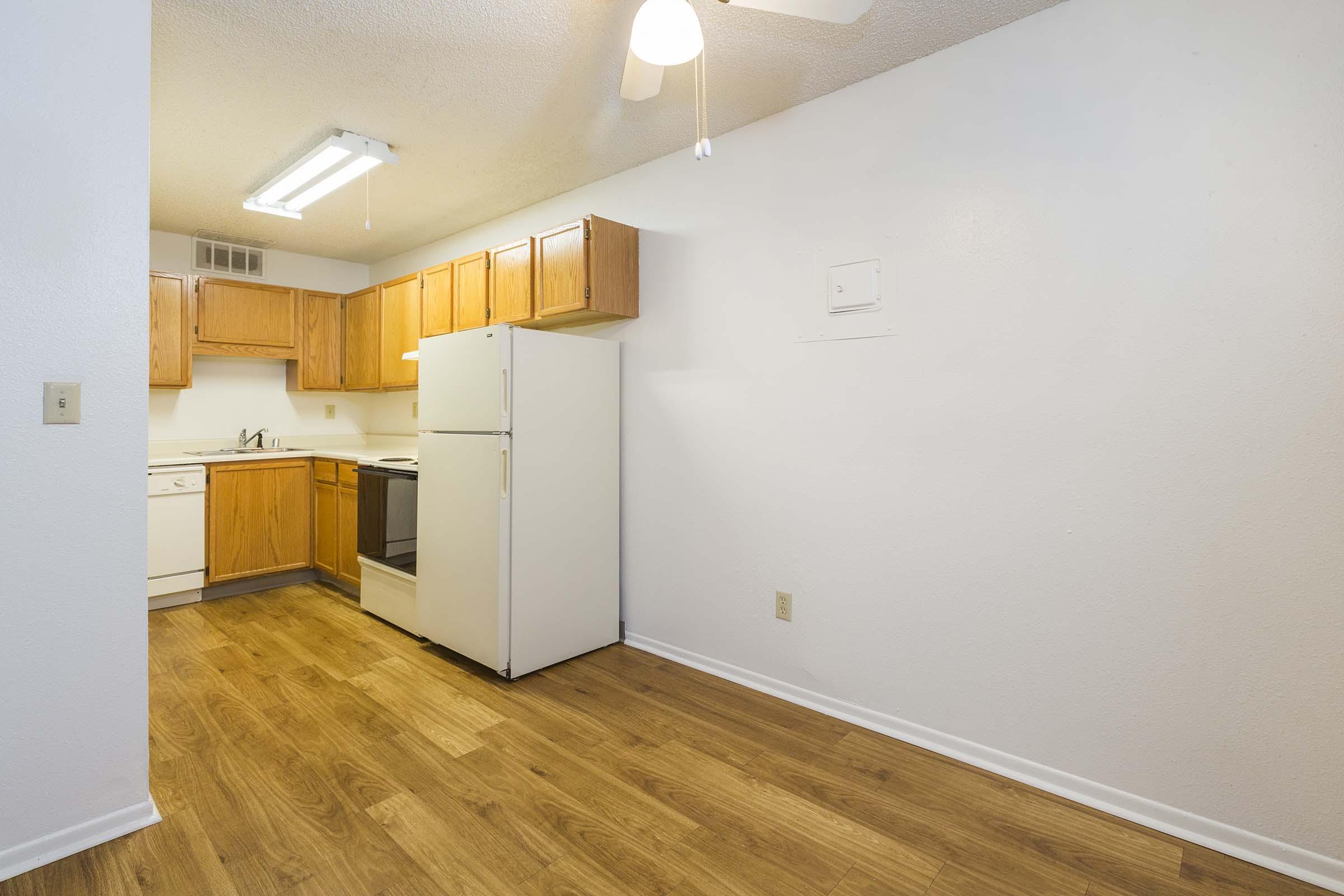 A small kitchen featuring wooden cabinets, a white refrigerator, an electric stove, and a sink. The space has hardwood flooring and a ceiling fan. There is a window providing natural light, enhancing the bright and clean atmosphere of the room.