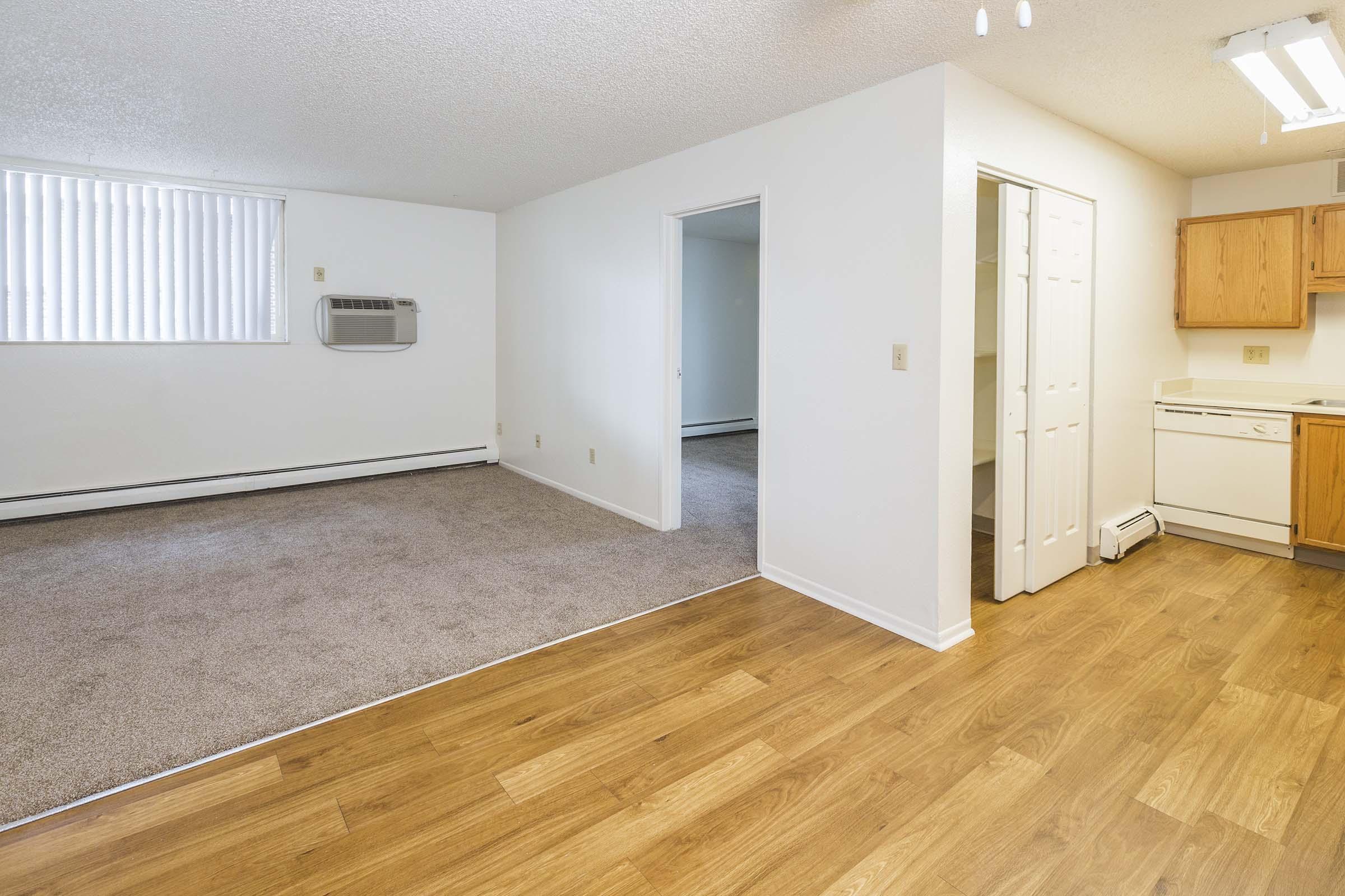 A spacious, neutral-colored living area with plush carpet and laminate flooring. There is a wall-mounted air conditioning unit in the corner, and a doorway leading to a kitchen with wooden cabinets and appliances. Natural light fills the room through large windows with vertical blinds.