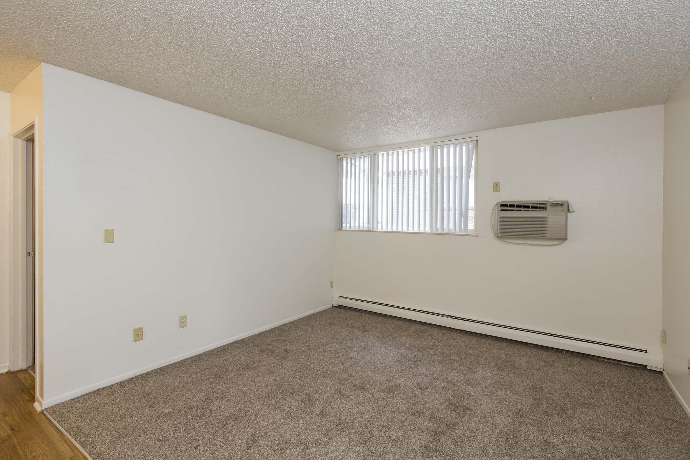 Empty living room with beige carpet and light-colored walls. A window on one side has vertical blinds, and an air conditioning unit is mounted below it. The space is well-lit and simple, with a door leading to another room on the left.