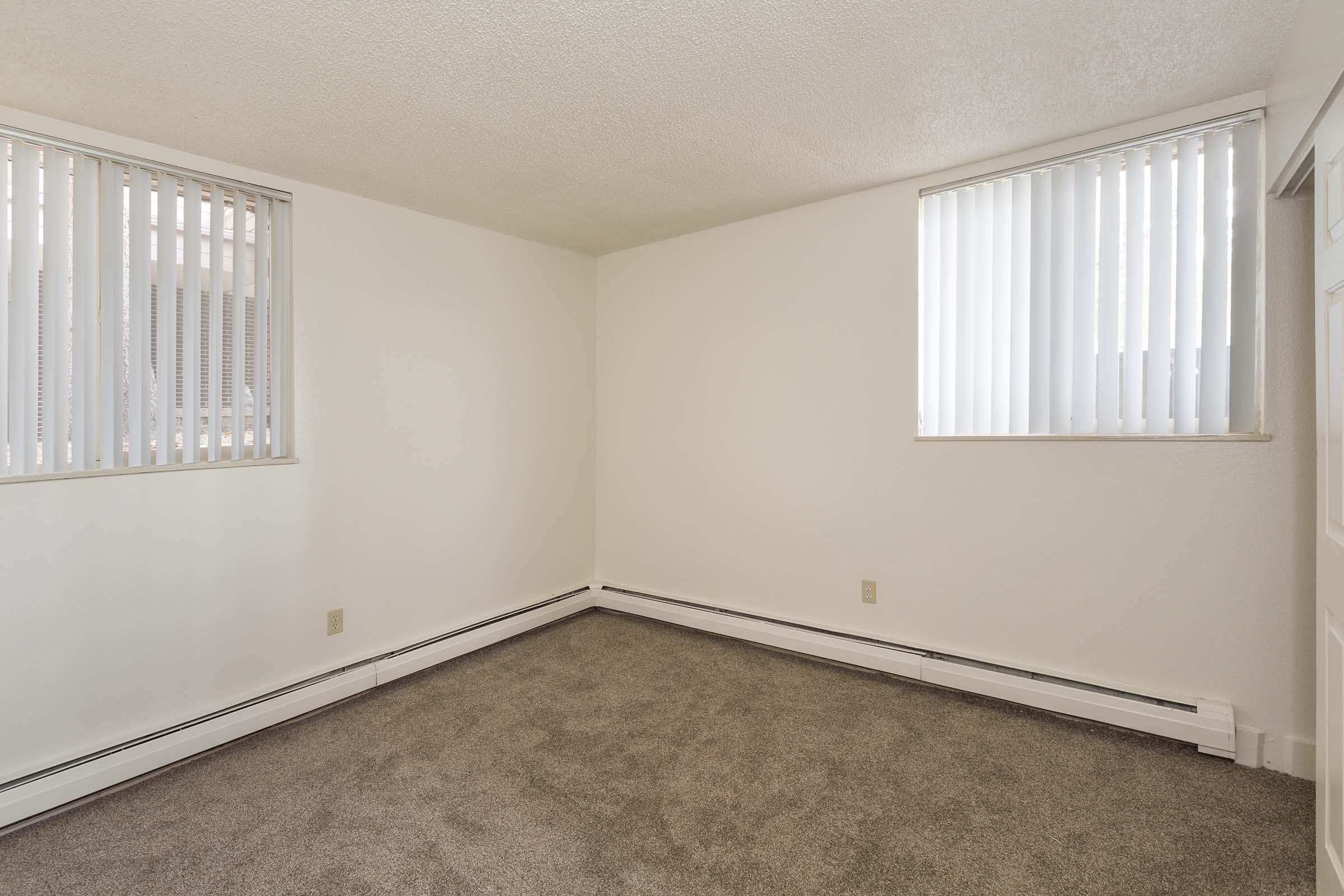 Empty room with beige carpet and light-colored walls. There are two windows with vertical blinds letting in natural light. Baseboard heating is visible along the walls, and the space appears clean and uncluttered, ready for furnishings.