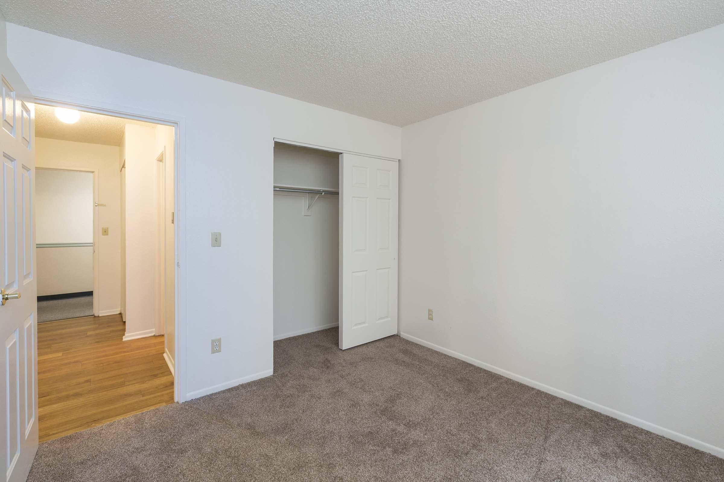 Empty room with beige carpet and white walls. A door on the left leads to a closet with a sliding door. There is another door in the background, suggesting access to a hallway. The lighting is bright, and the space appears clean and well-maintained.