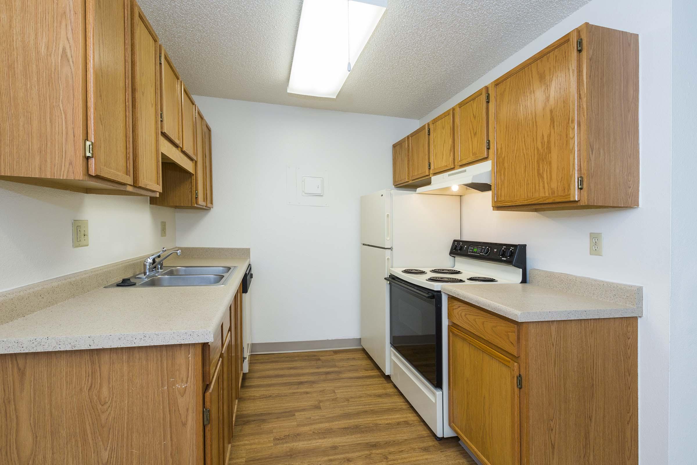 A small kitchen featuring light wood cabinets, a stainless steel sink, a white refrigerator, and an electric stove with an oven. The countertops are beige, and the floor has a wood-like appearance. A ceiling light provides illumination, and the walls are painted white.