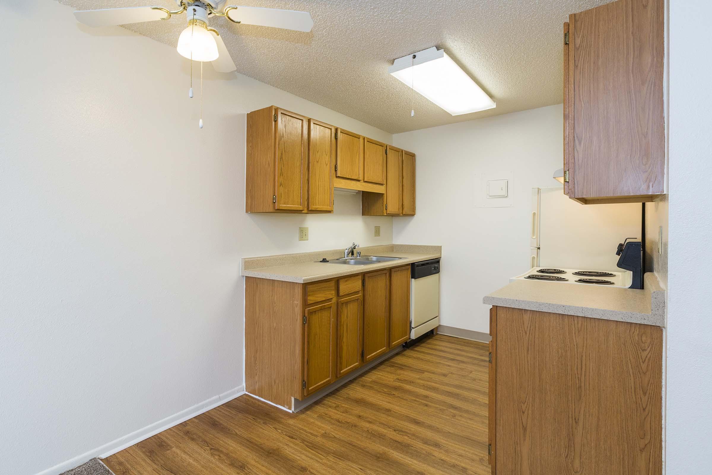 A small kitchen with wooden cabinets, a countertop, and appliances, including a dishwasher and a stove. The room features a ceiling fan, a light fixture, and a windowless wall. The floor is covered with wood-like laminate, creating a warm and inviting atmosphere.