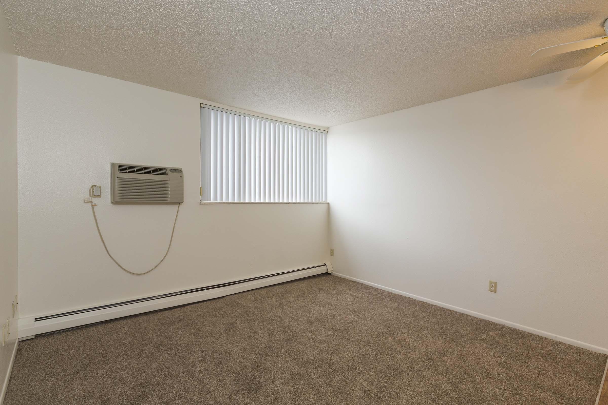 A vacant room featuring light beige carpet and white walls. A window with vertical blinds provides natural light, and an air conditioning unit is mounted on the wall. The space is minimalistic and uncluttered, ideal for personalized decor or furnishings.