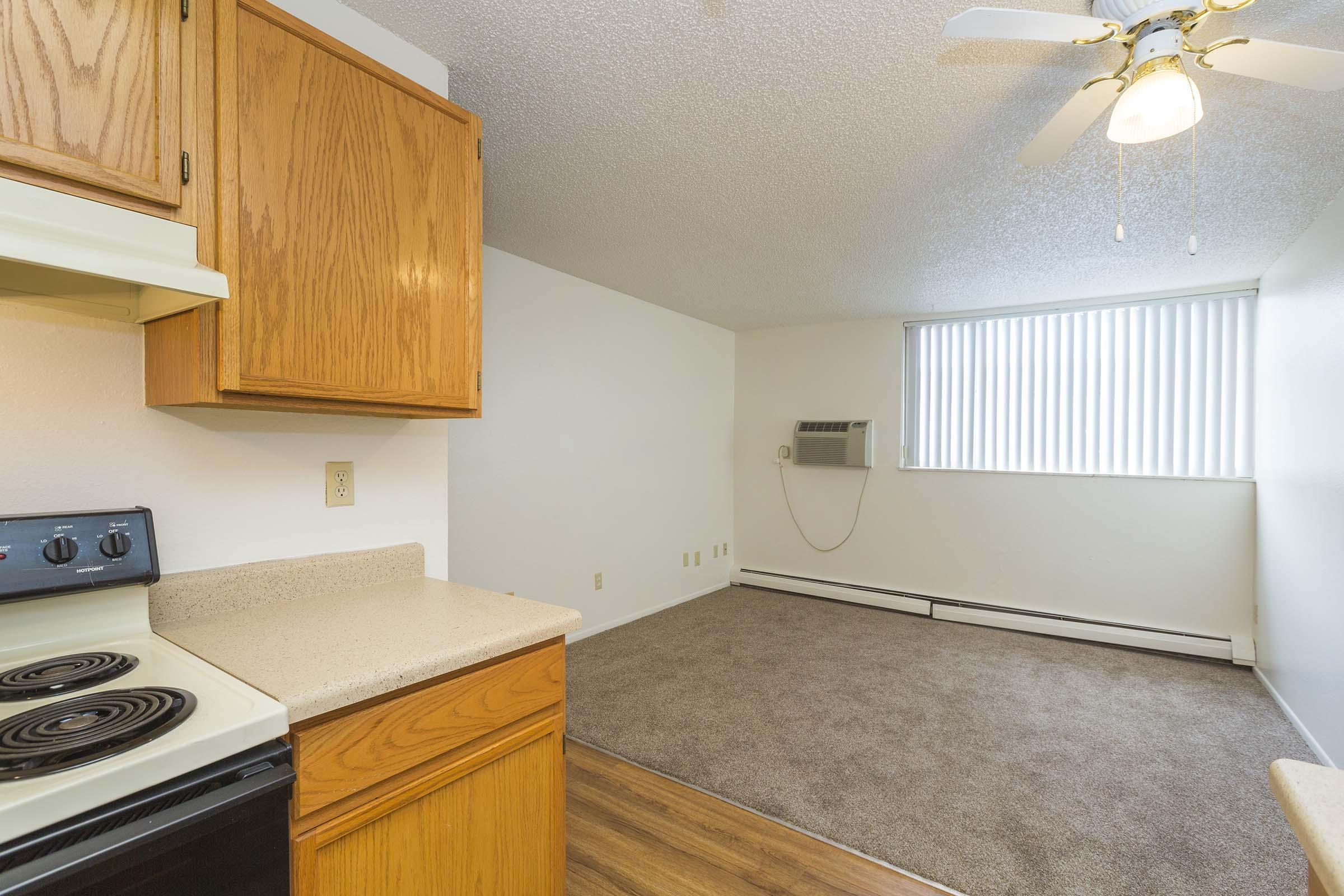 A small kitchen area featuring wooden cabinetry, a stove, and a countertop, leading to a carpeted living space. There is a window with vertical blinds allowing natural light, and an air conditioning unit on the wall. The overall color scheme is neutral and inviting.