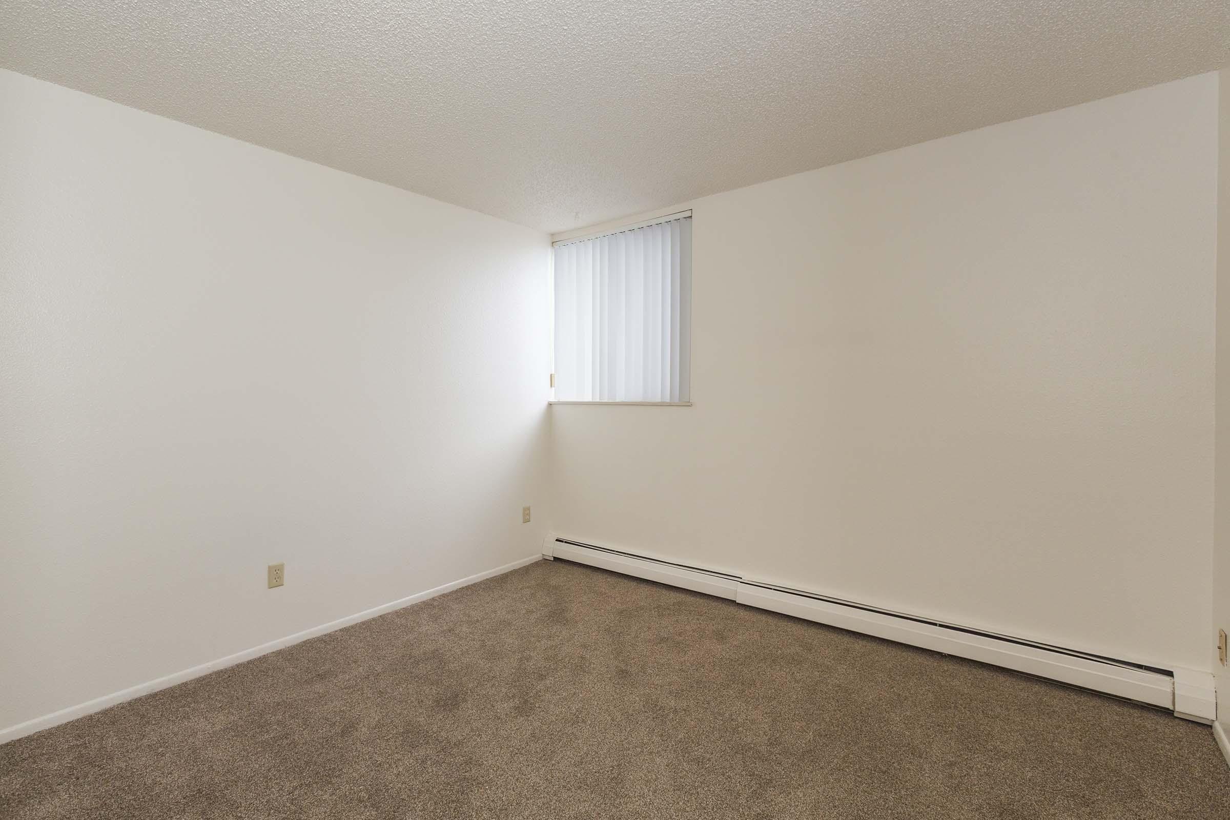 A small, empty room with beige carpet and white walls. There is a single window on one wall with vertical blinds partially opened, allowing light to enter. The room appears clean and unfurnished, providing a blank canvas for potential decoration or use.
