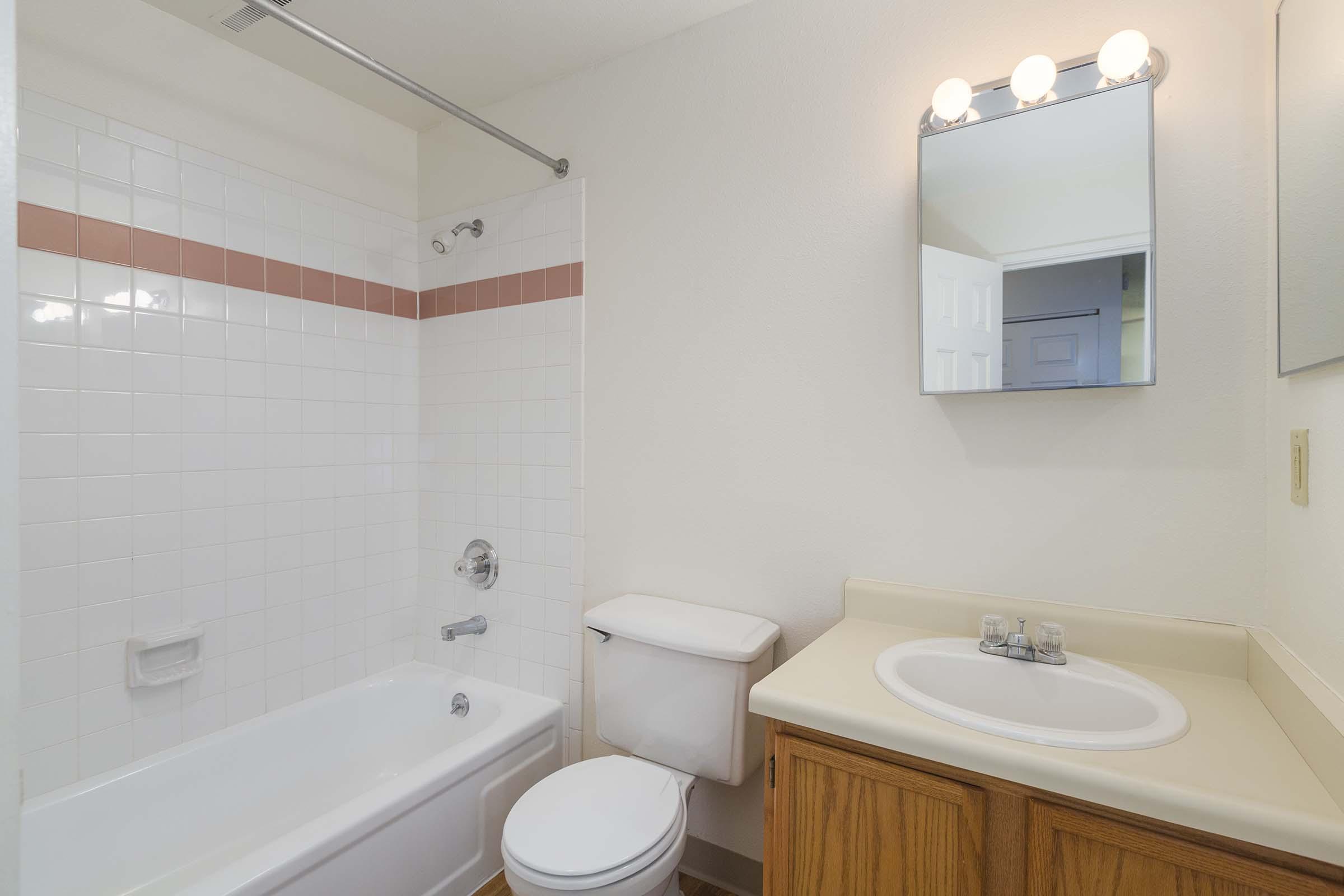 A tidy and well-lit bathroom featuring a bathtub with a shower, a toilet, a mirror above the sink, and a wooden vanity. White tiles with a pink accent line the walls, and there is a small window visible in the background.