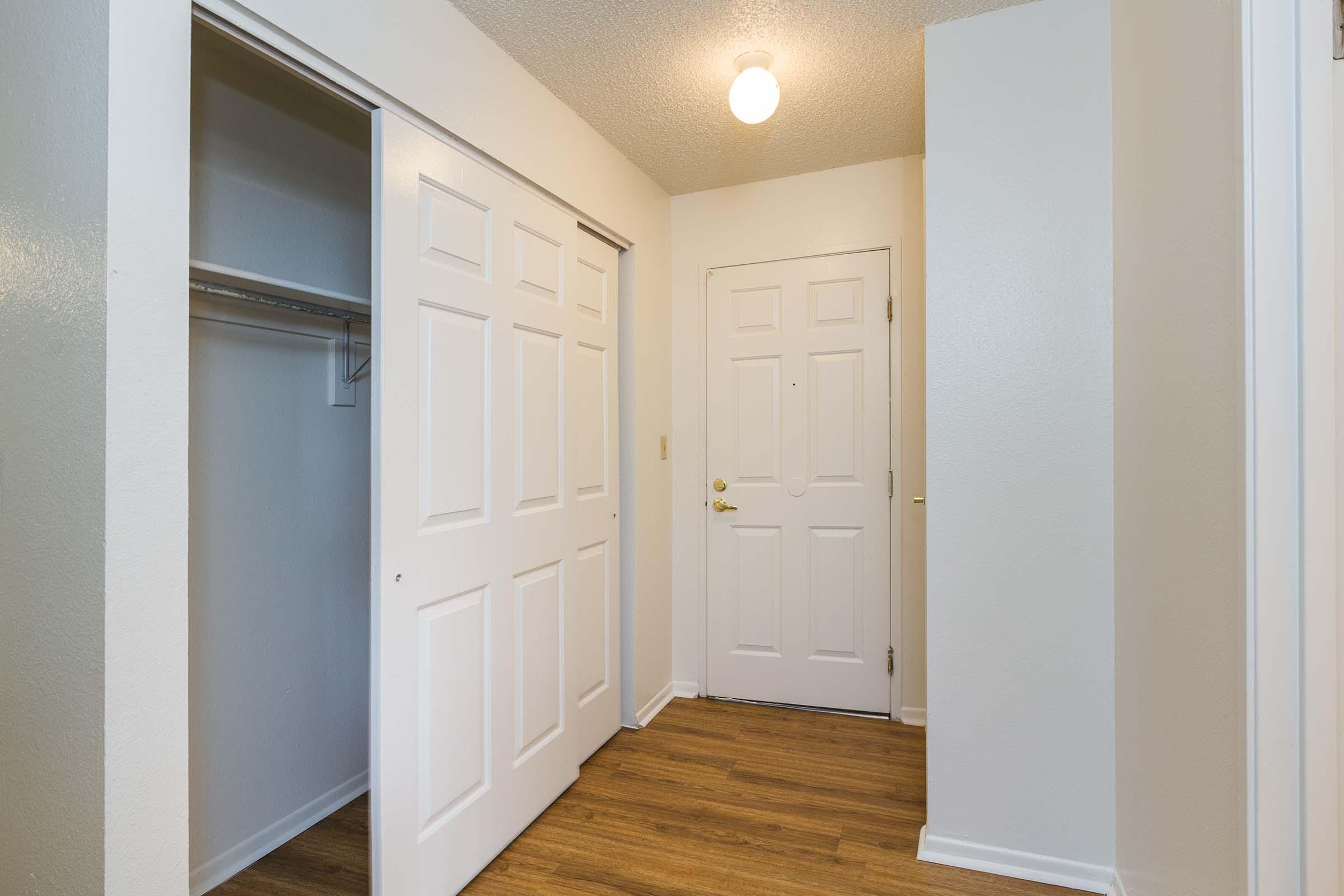 A well-lit entryway featuring a closed door with a doorknob and a small window, adjacent to a closet with sliding white doors. The space has a light-colored wall and a light wooden floor, creating a welcoming atmosphere.