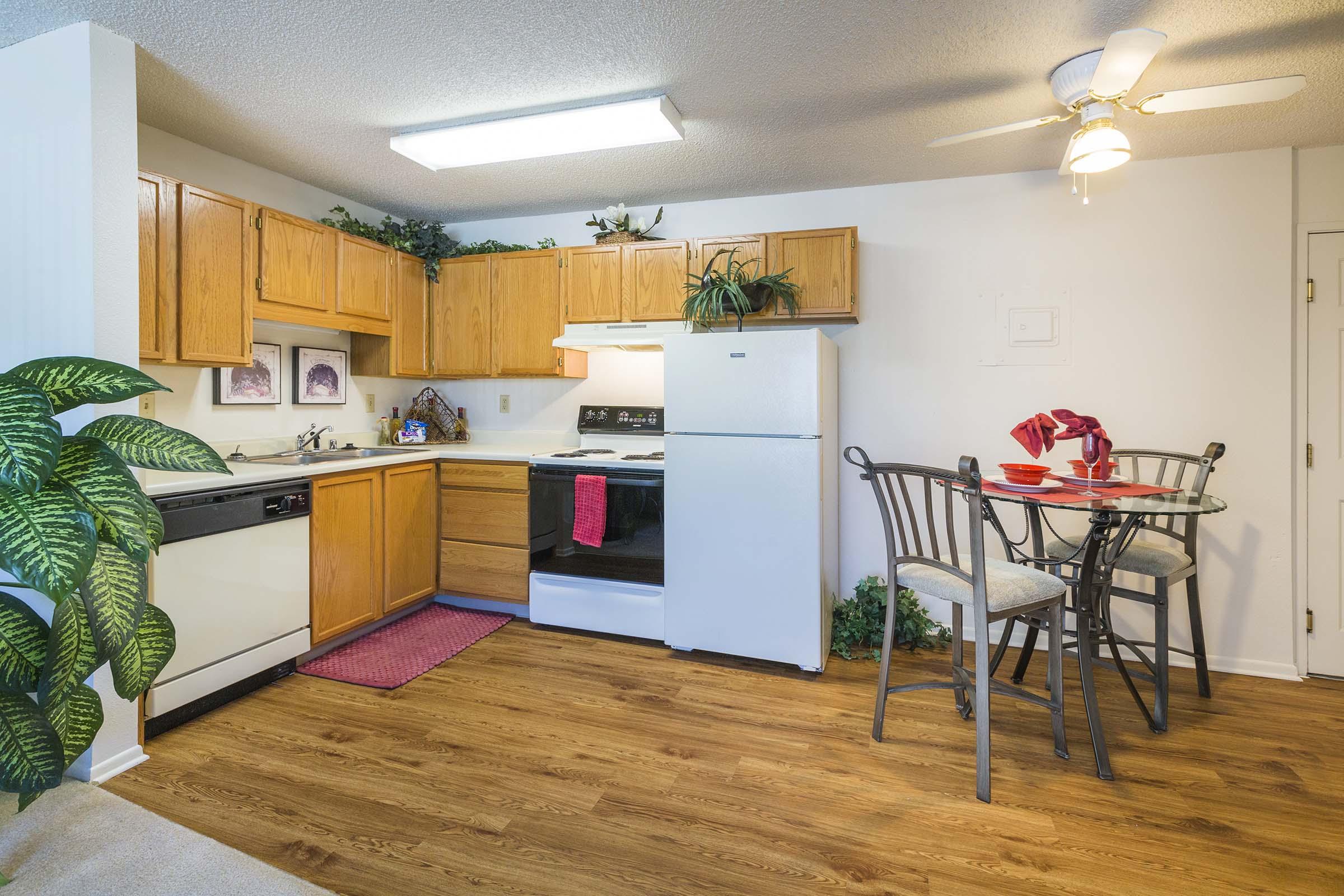 A cozy kitchen featuring wooden cabinets, a white refrigerator, and an electric stove. A round dining table set for two sits beside a small window, with chairs and a red table setting. Green plants adorn the space, adding a touch of greenery to the warm, inviting atmosphere.