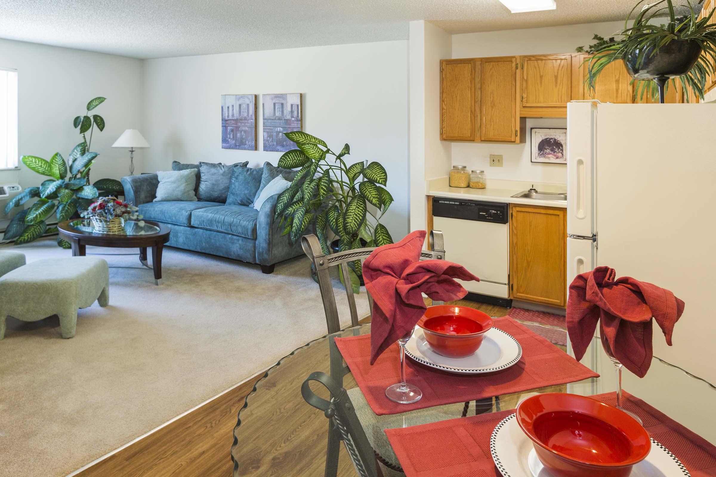 A cozy living space featuring a light-filled room with a teal sofa, a round wooden coffee table, and decorative plants. The adjacent kitchen includes wooden cabinetry, white appliances, and a dining area set with red table settings, including bowls and napkins, creating an inviting atmosphere.