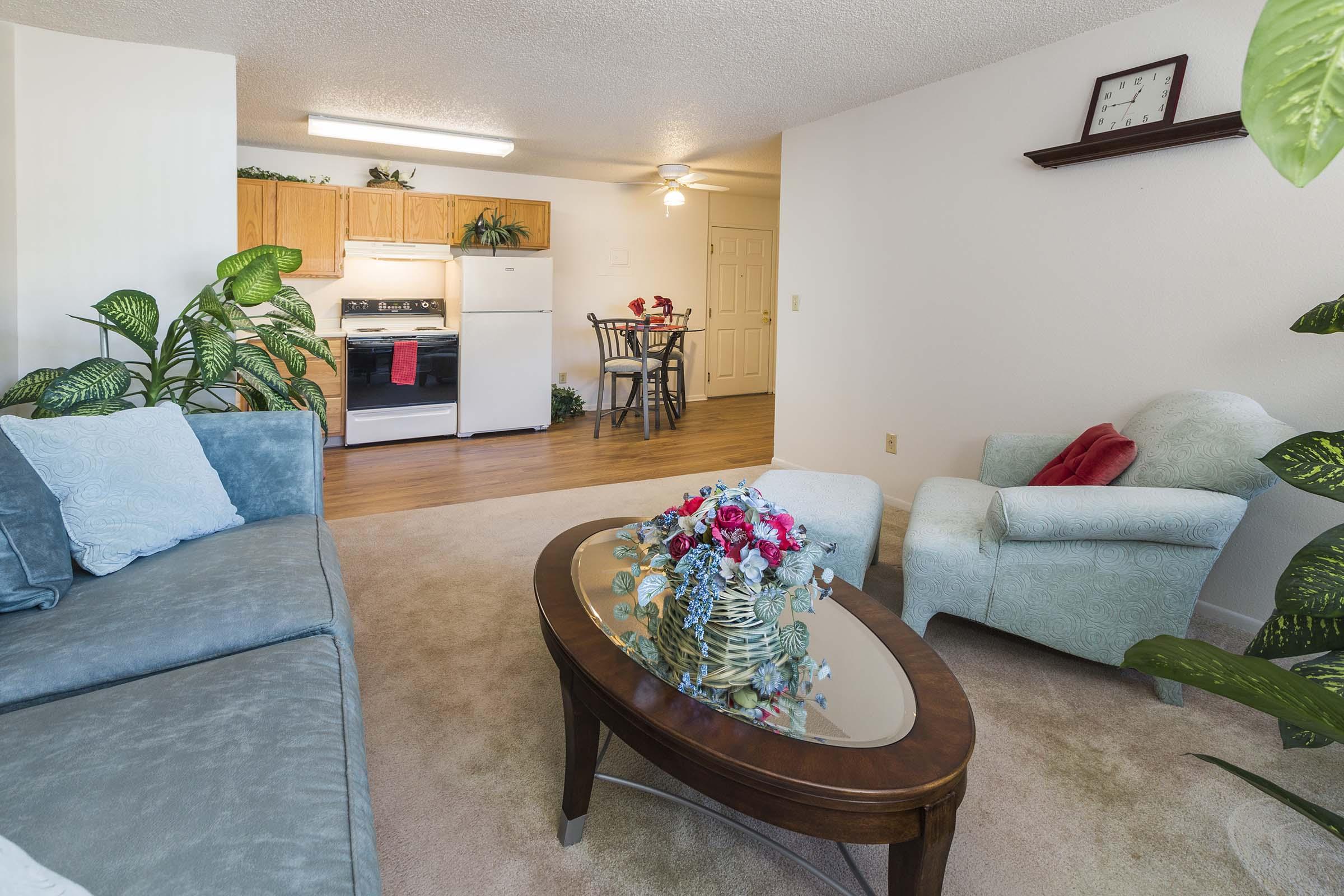 A cozy living area featuring a light blue sofa, a glass coffee table with a floral centerpiece, and a light green armchair. In the background, there's a small dining table and kitchen with wooden cabinets, a stove, and a refrigerator. The space is adorned with indoor plants and has soft lighting.