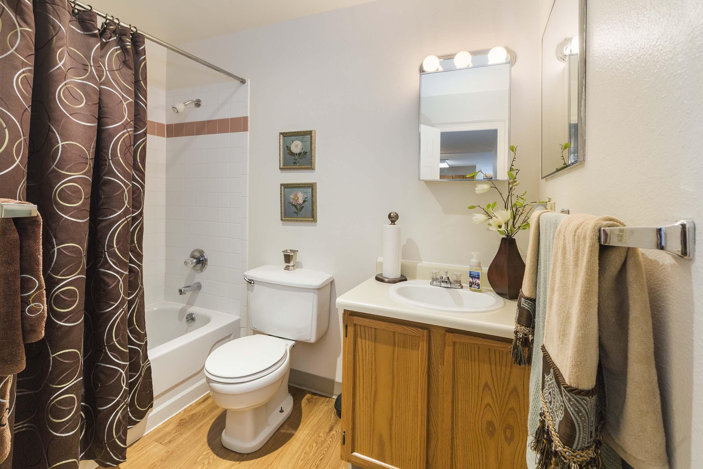 A well-lit bathroom featuring a bathtub with a shower, a toilet, and a wooden vanity. The vanity includes a sink with toiletries and decorative accents. A dark brown shower curtain adds contrast, while framed artwork and a small vase with flowers enhance the decor. Softwood flooring completes the space.