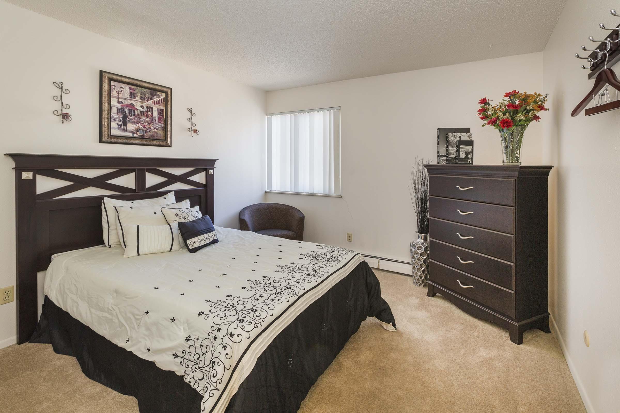 A cozy bedroom featuring a queen-sized bed with a black and white patterned comforter. There is a framed artwork on the wall, a brown armchair in the corner, and a dark wooden dresser with decorative items. Natural light comes from a window with vertical blinds. Soft beige carpet underfoot.