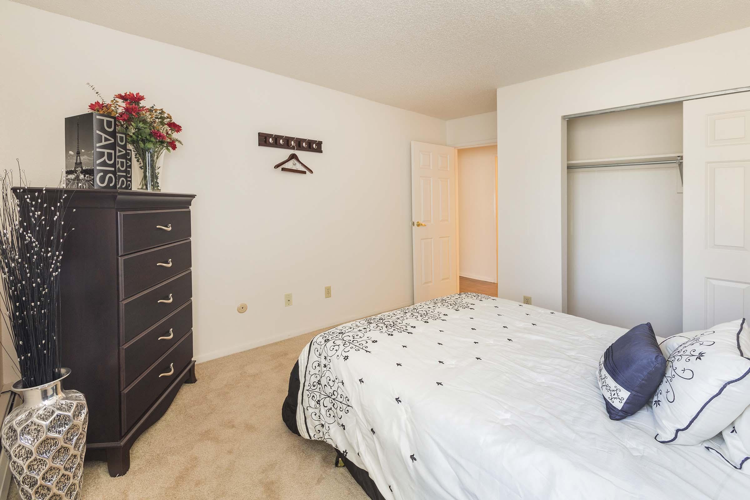 A bright bedroom featuring a neatly made bed with a white comforter and decorative pillows. A dark wood dresser stands against one wall, adorned with a vase of flowers and a decorative item labeled "Paris." There is a closet with white folding doors, and light-colored walls create a spacious feel.