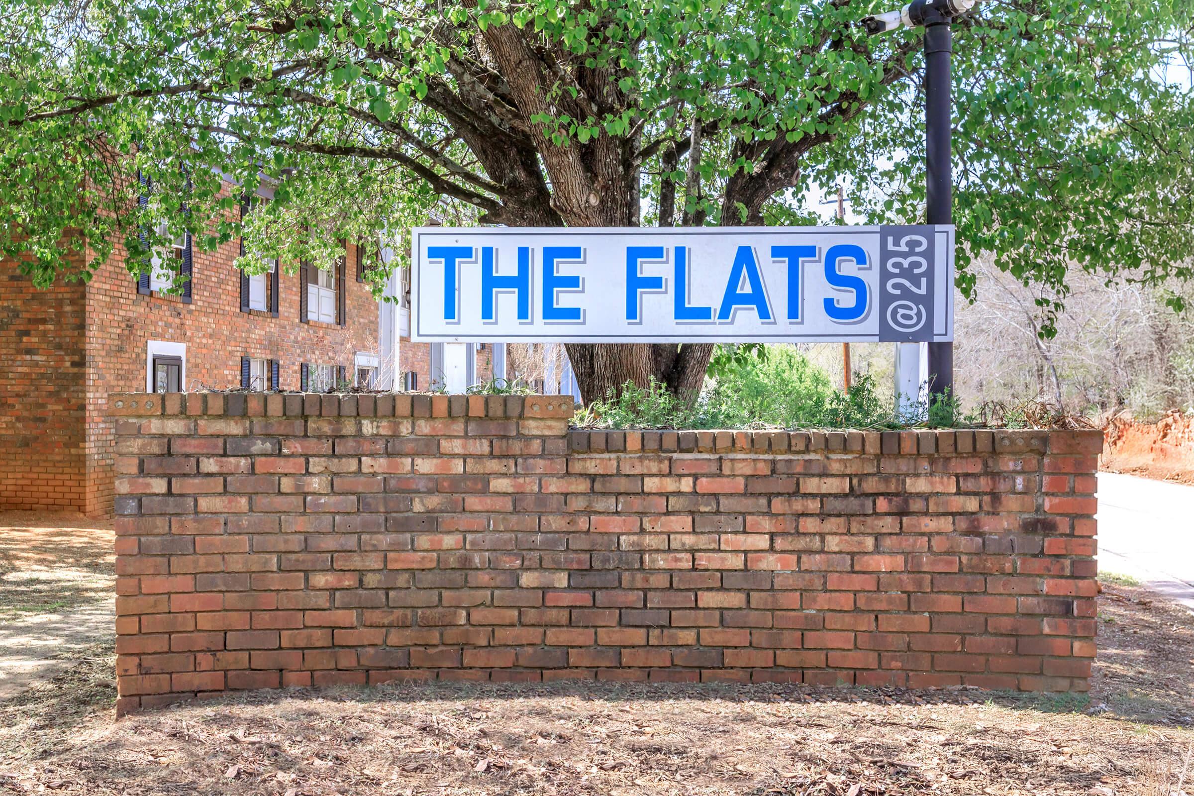Sign for "The Flats" at 235, displayed on a brick wall with greenery in the background. The sign features large blue letters on a white background, indicating the location. The surrounding area appears to be a residential complex with trees and grass.