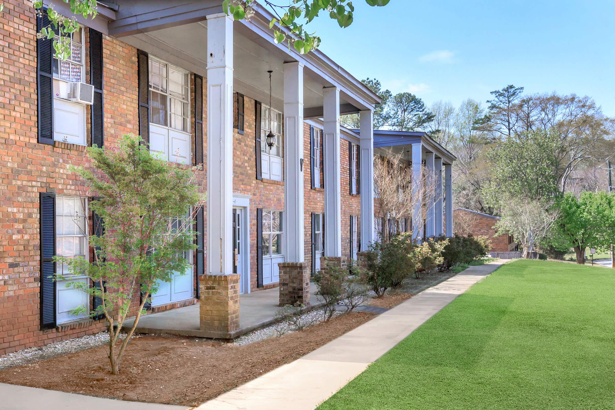 A brick building with multiple entrances featuring white doors and large windows. The structure is flanked by green shrubs and trees, with a sidewalk leading up to it. The scene includes lush grass and a clear blue sky in the background.
