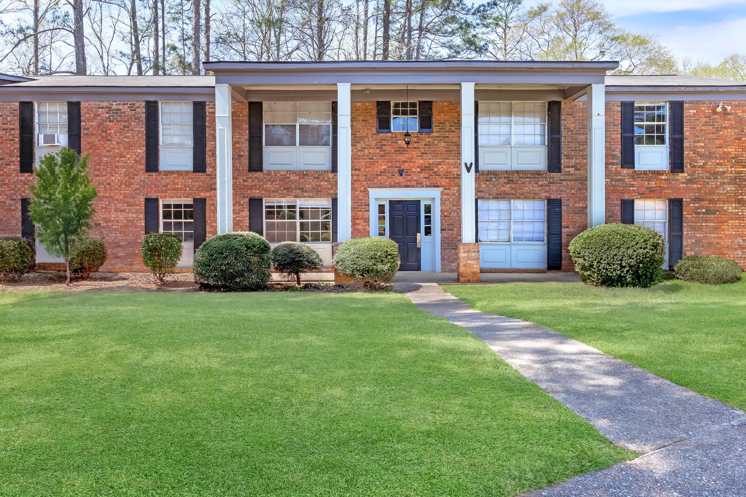 A brick apartment building with a symmetrical facade featuring large windows, a central entrance, and decorative columns. The surrounding area has well-maintained lawns and shrubs, with a pathway leading to the front door. Trees are visible in the background, indicating a serene environment.