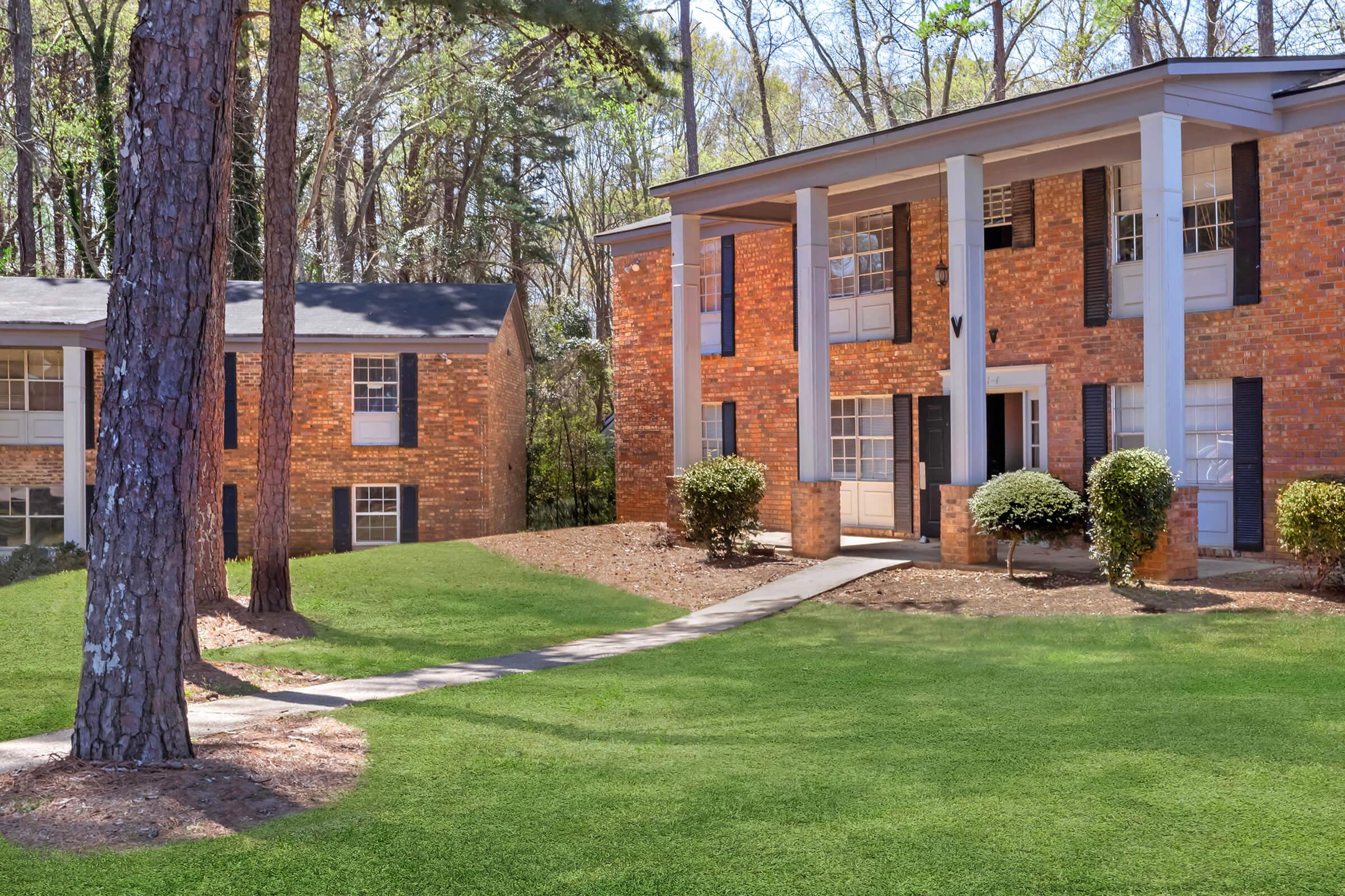 Two brick apartment buildings surrounded by green grass and trees. One building has a white-columned entrance with a pathway leading to it, while the other is visible in the background with multiple windows. The setting is bright and sunny, featuring landscaped shrubs.
