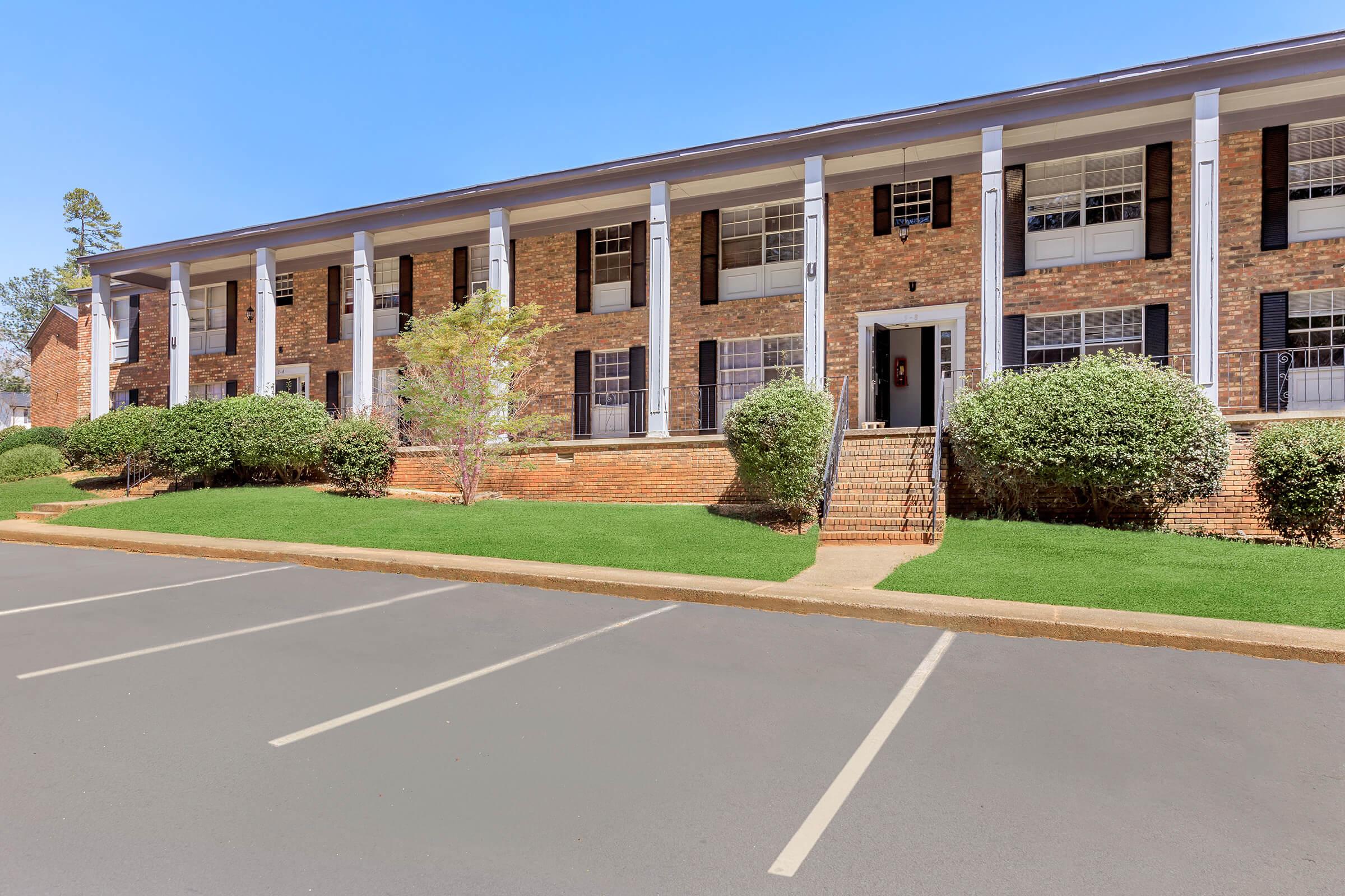 A brick apartment building featuring two stories, multiple windows with black shutters, and a landscaped entrance. Green shrubs line the front, and a set of steps leads to the main door. The parking area in front is empty, with clear blue skies in the background.