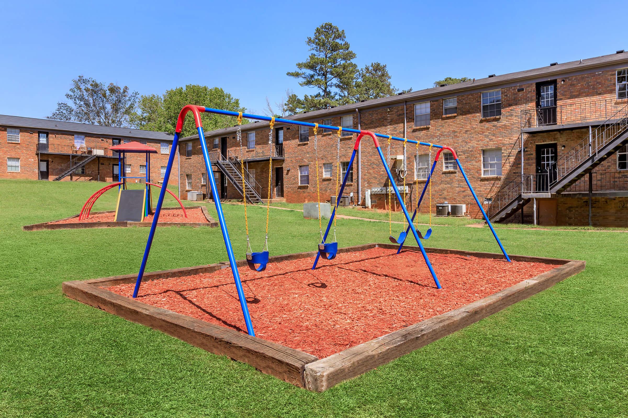 A colorful playground featuring blue and red swings on a bed of mulch, set in a grassy area in front of brick apartment buildings. The scene is bright and inviting, with clear blue skies above and a few trees in the background, making it a cheerful outdoor space for children.