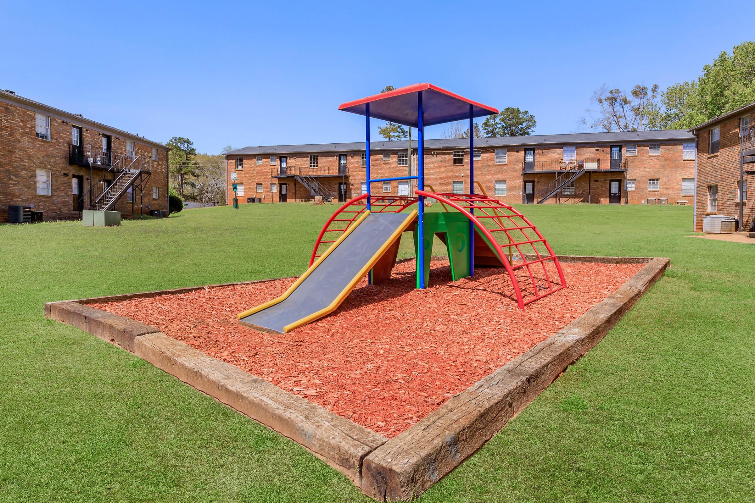 A colorful playground structure featuring a slide, set on a bed of mulch, surrounded by grassy areas. In the background, there are brick apartment buildings under a clear blue sky.