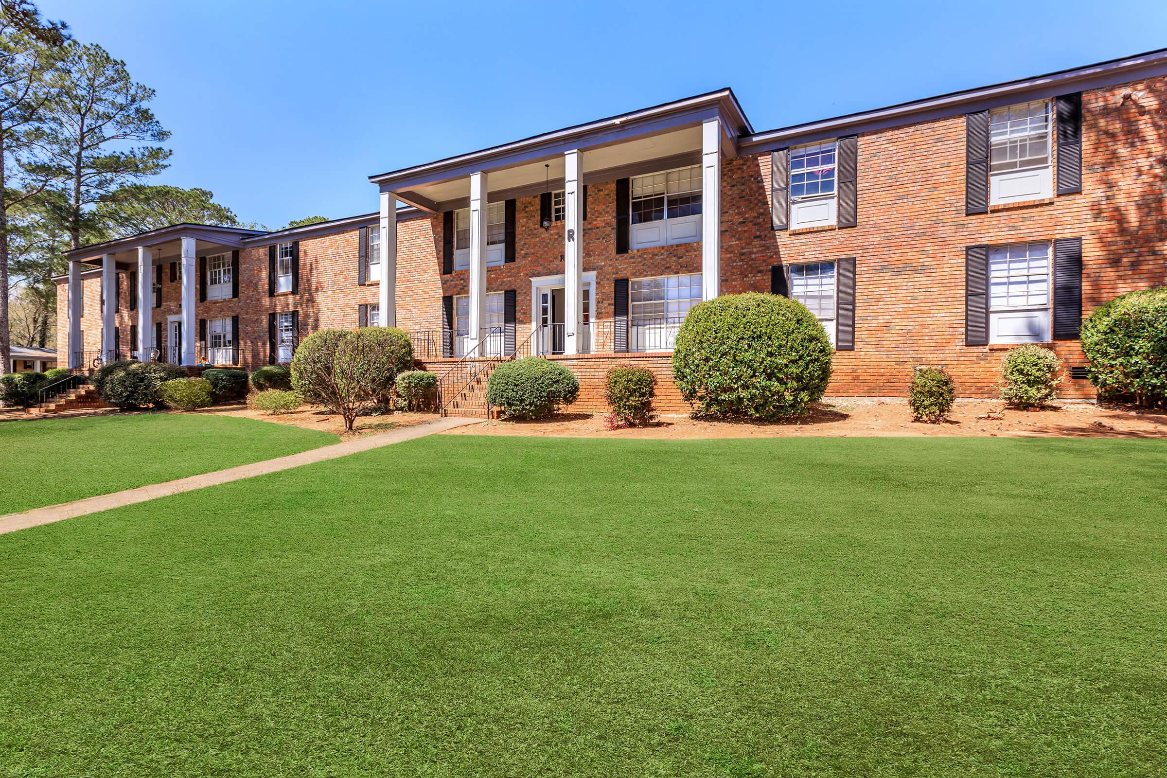 Two-story brick apartment building surrounded by manicured landscaping, featuring a symmetrical facade with multiple entrances, large windows, and a well-maintained lawn. The sunny blue sky adds to the inviting atmosphere of the residential area.