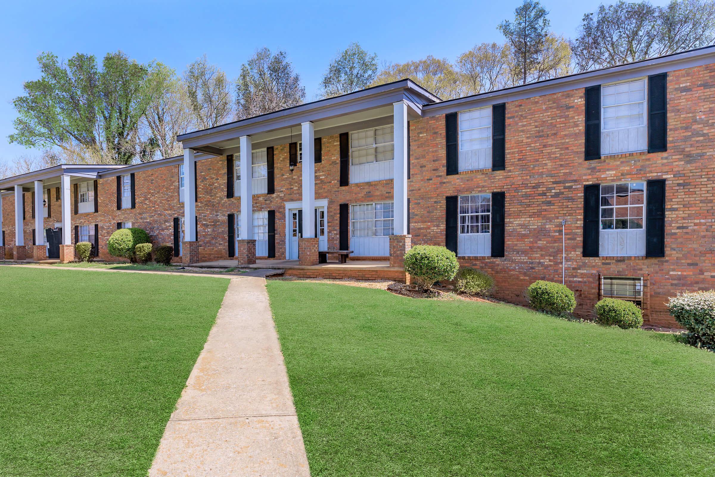 Exterior view of a brick apartment building with multiple windows and a well-maintained front lawn. The structure features white columns at the entrance and dark shutters on the windows. Surrounding greenery includes trimmed bushes and trees in the background under a clear blue sky.