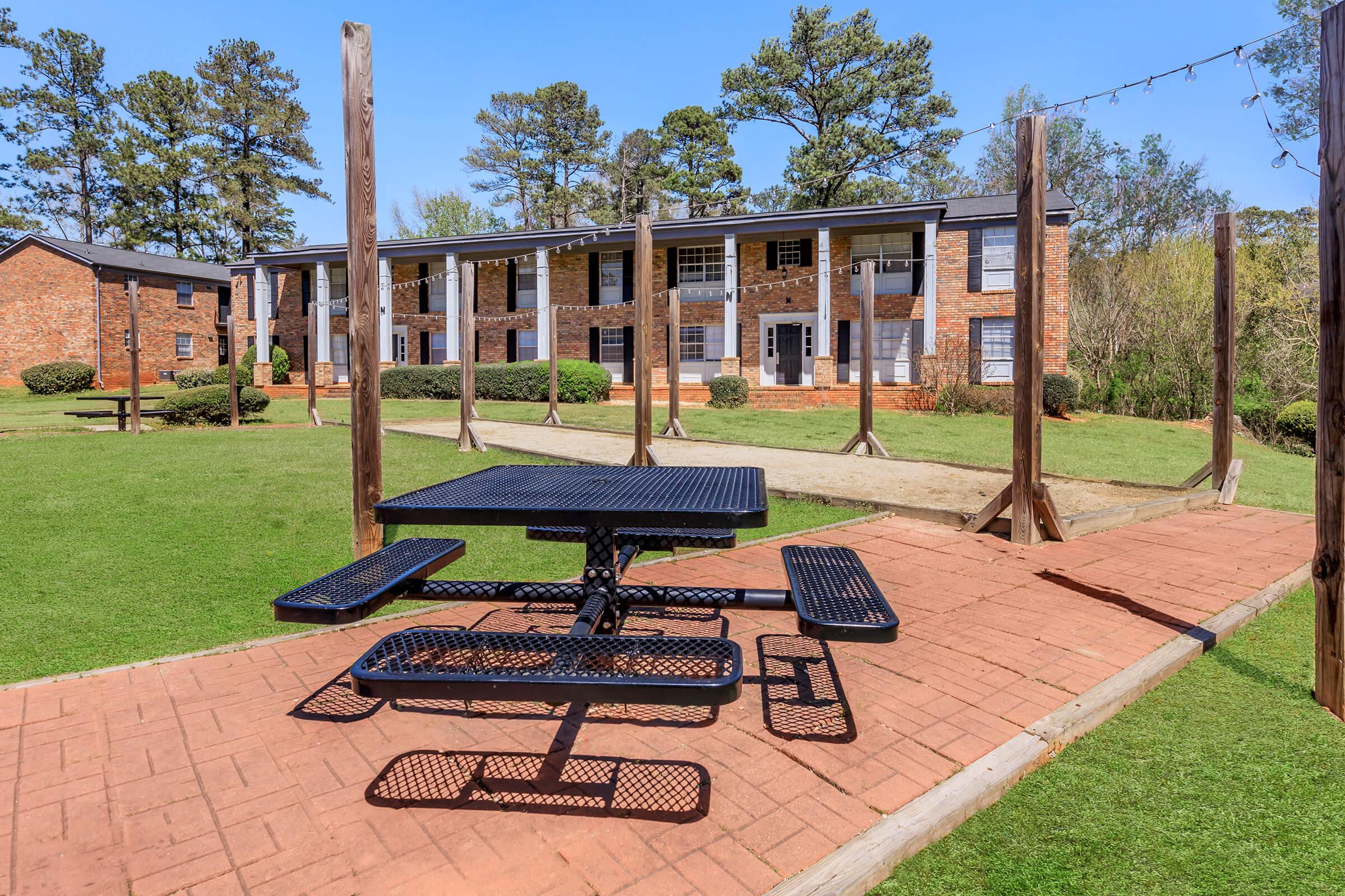 A picnic table with attached benches sits on a brick patio surrounded by green grass. In the background, there are two-story brick apartment buildings with balconies, framed by trees under a clear blue sky. Some wooden posts suggest possible hanging lights.