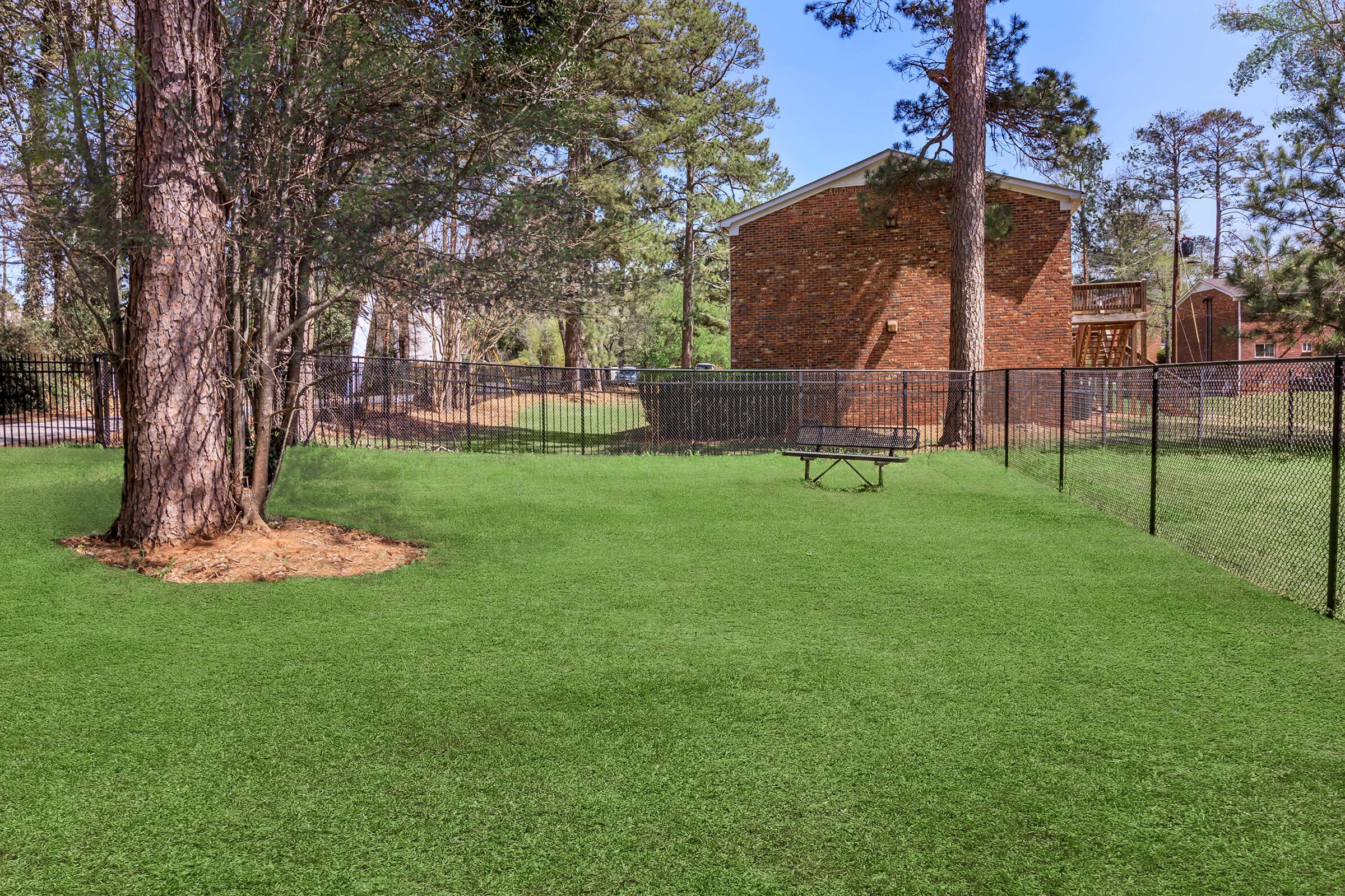 A grassy area enclosed by a black fence, featuring a tree and a bench. In the background, there is a brick building and several trees, creating a serene outdoor space. The blue sky adds to the peaceful atmosphere.