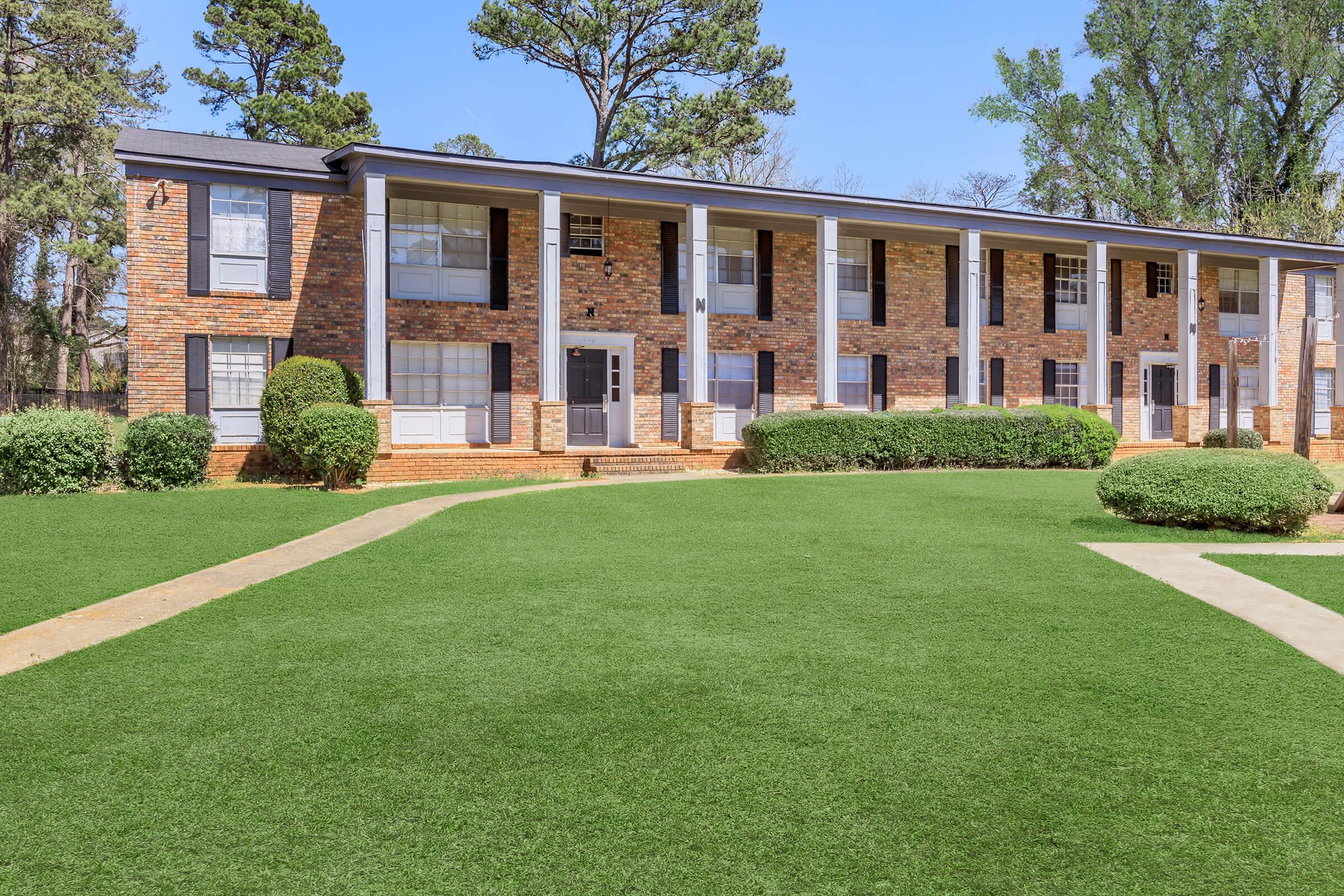 A well-maintained two-story apartment building with a brick exterior, featuring multiple units and large windows. The manicured lawn is lush and green, bordered by neatly trimmed bushes. A paved walkway leads to the entrance, and tall trees provide shade in the background under a clear blue sky.