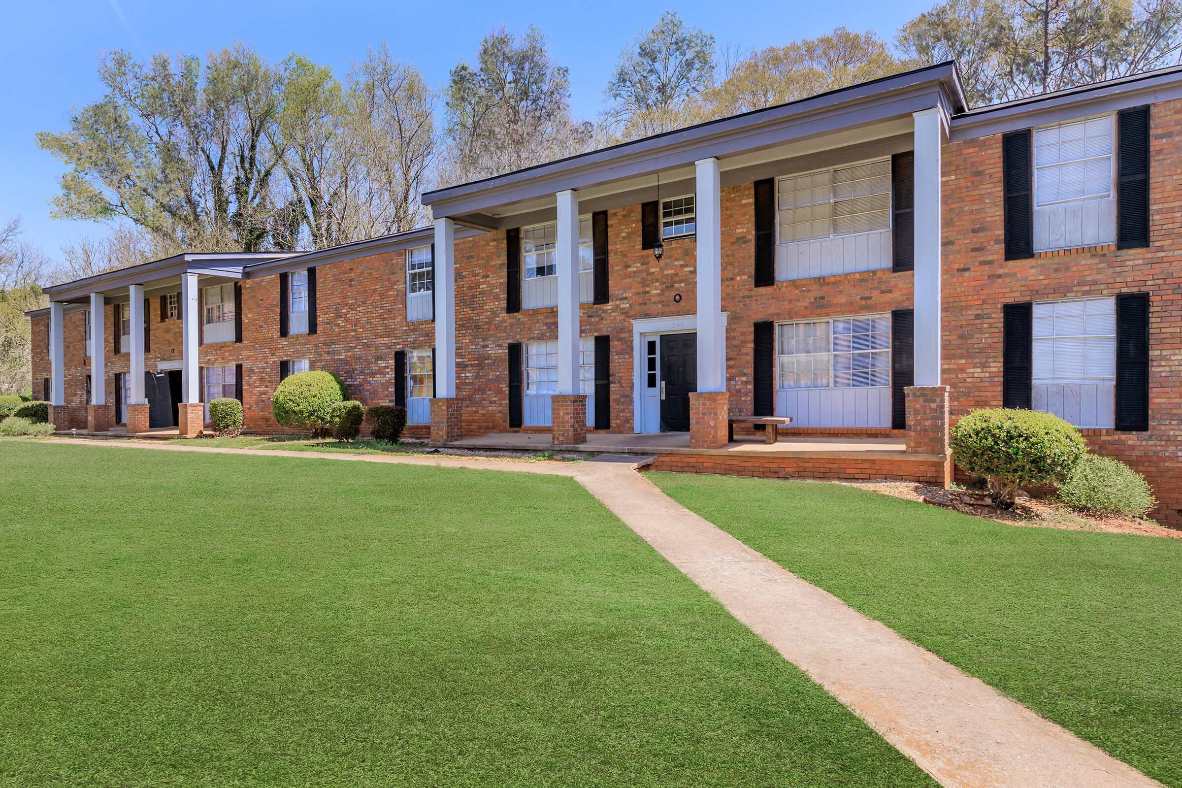 A brick apartment building with multiple entrances and large windows, surrounded by manicured lawns and bushes. The building has two stories and a pathway leading to the main entrance, with trees in the background under a clear blue sky.