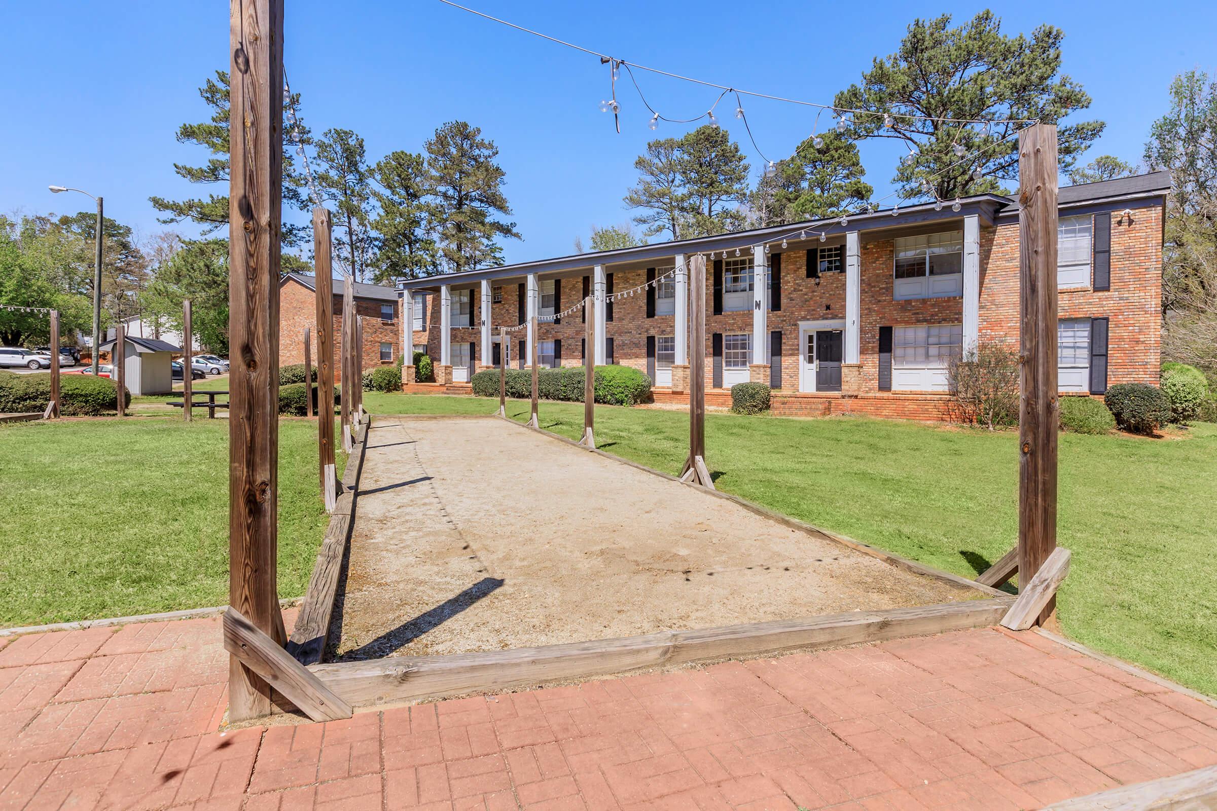 A view of an outdoor area featuring a gravel surface for playing games, surrounded by wooden posts. In the background, there are two-story brick apartment buildings with green lawns and trees. The sky is clear and blue, indicating a sunny day.