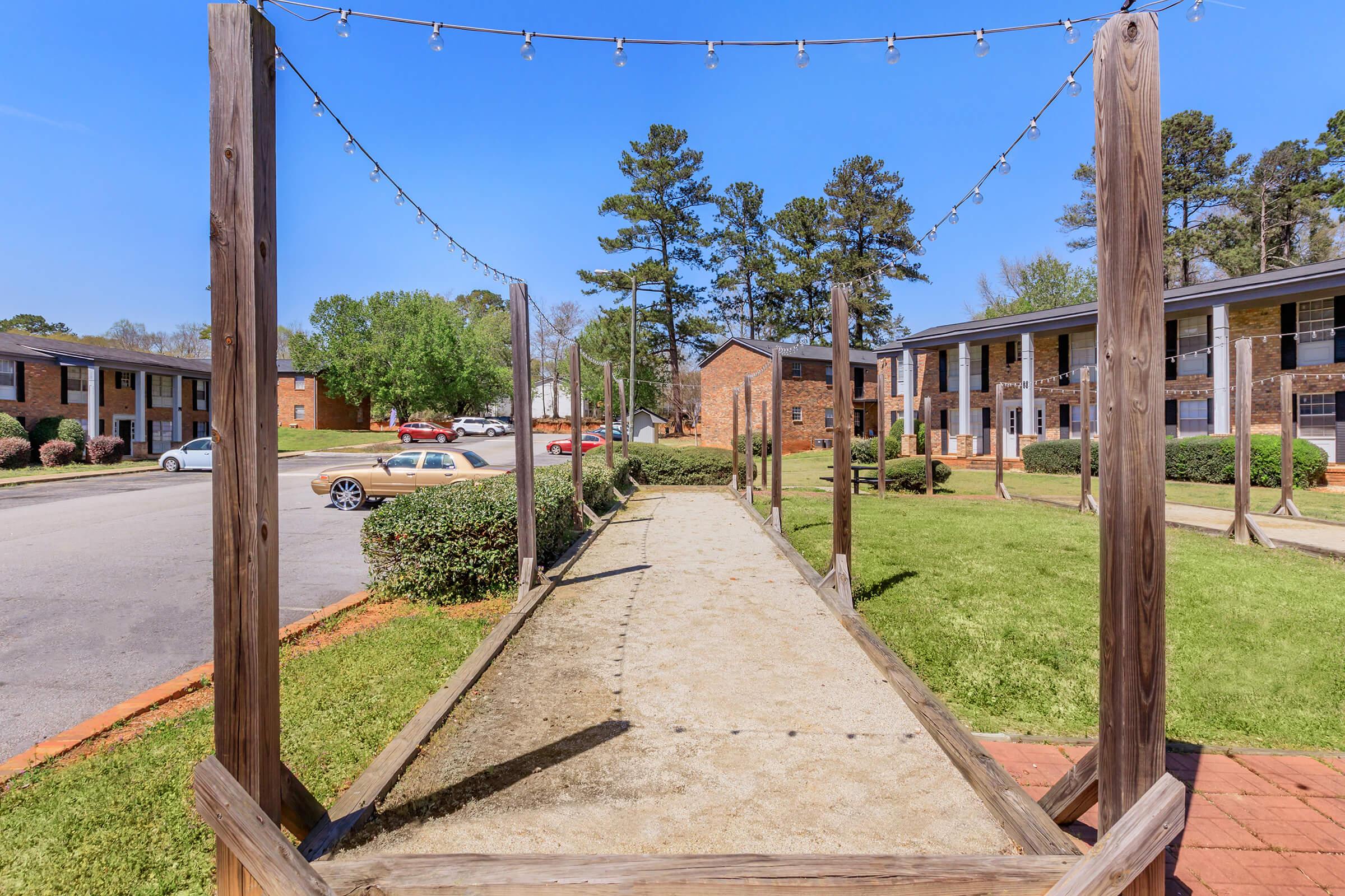 A walkway lined with wooden posts, leading through a grassy area towards residential buildings. The path is illuminated by string lights overhead. In the background, there are parked cars and a clear blue sky, surrounded by trees.