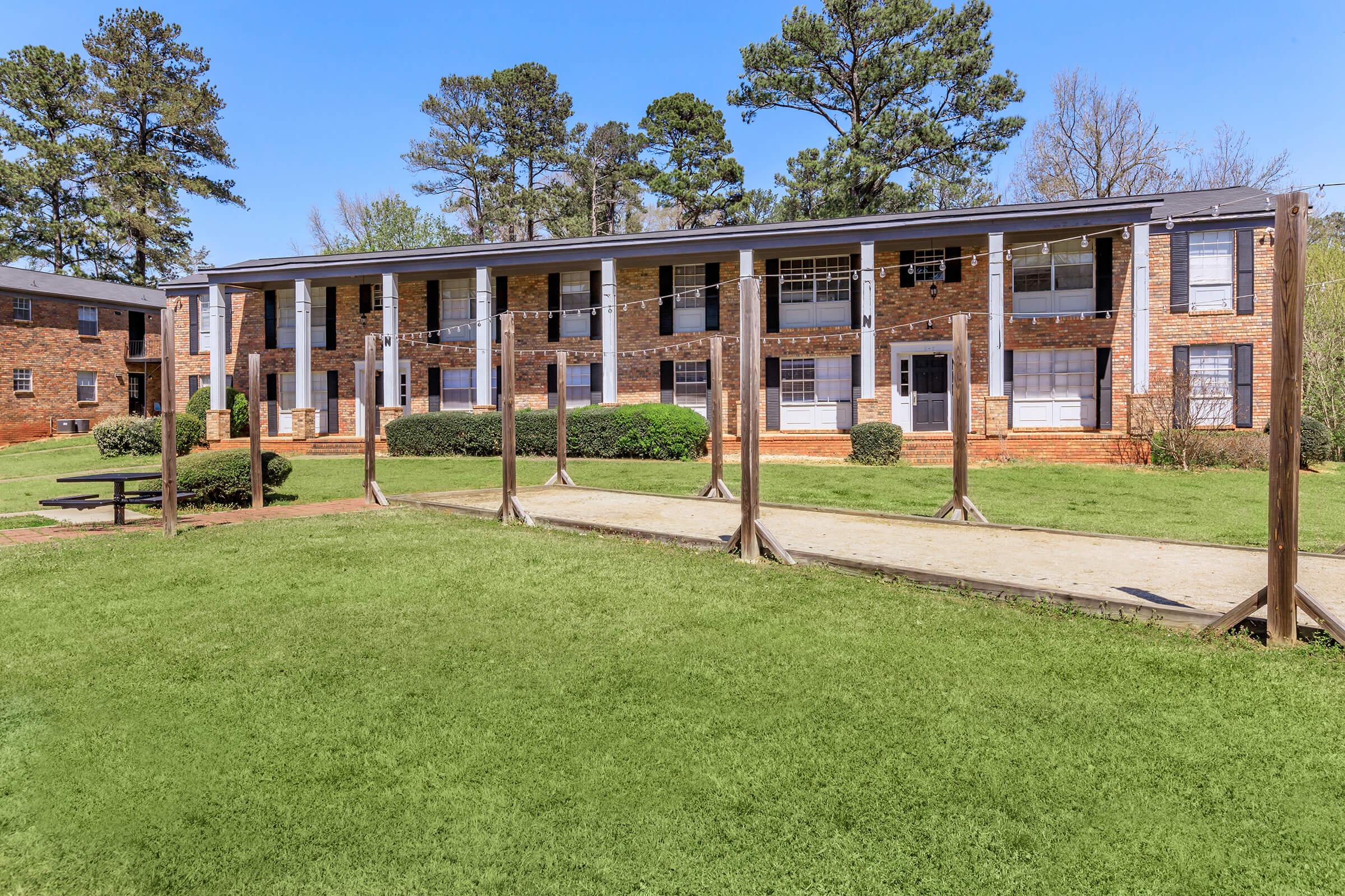 Two-story brick apartment building surrounded by green grass and trees. The building features several balconies, with multiple windows visible. In the foreground, there is a walkway leading to the entrance, along with picnic tables and clotheslines. Clear blue sky enhances the inviting atmosphere of the scene.