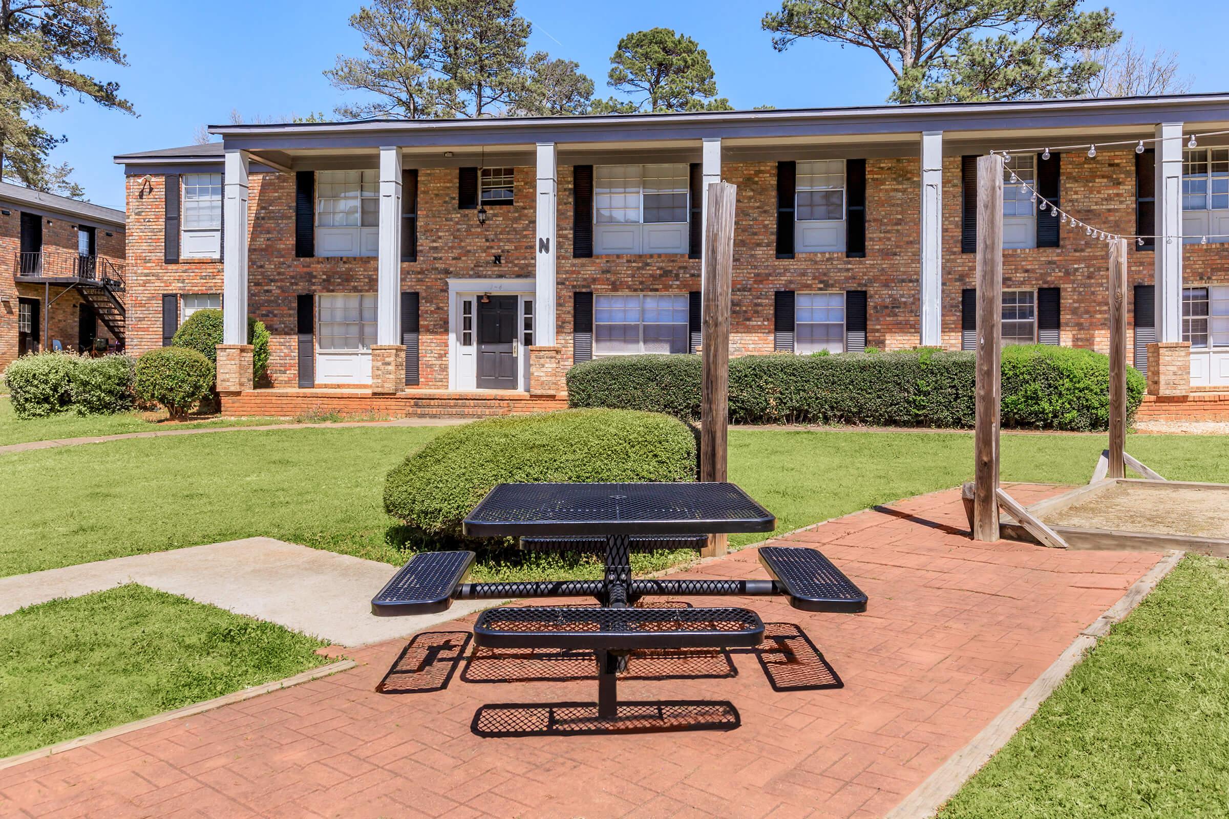 A brick apartment building with two levels, featuring multiple windows and a central entrance. In the foreground, there's a black picnic table on a paved pathway, surrounded by neatly trimmed grass and shrubbery. The background shows trees and a clear blue sky.