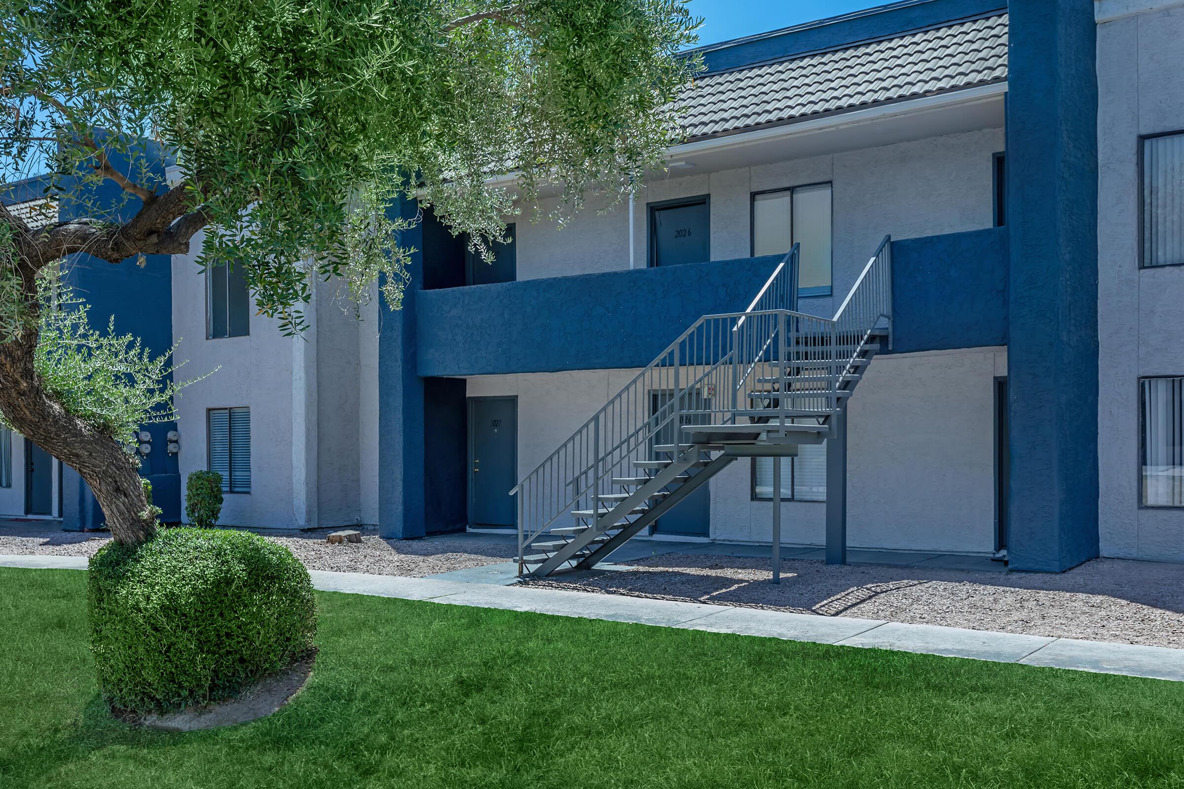 Blue and grey exterior apartment building tucked behind grass and sidewalk walkway
