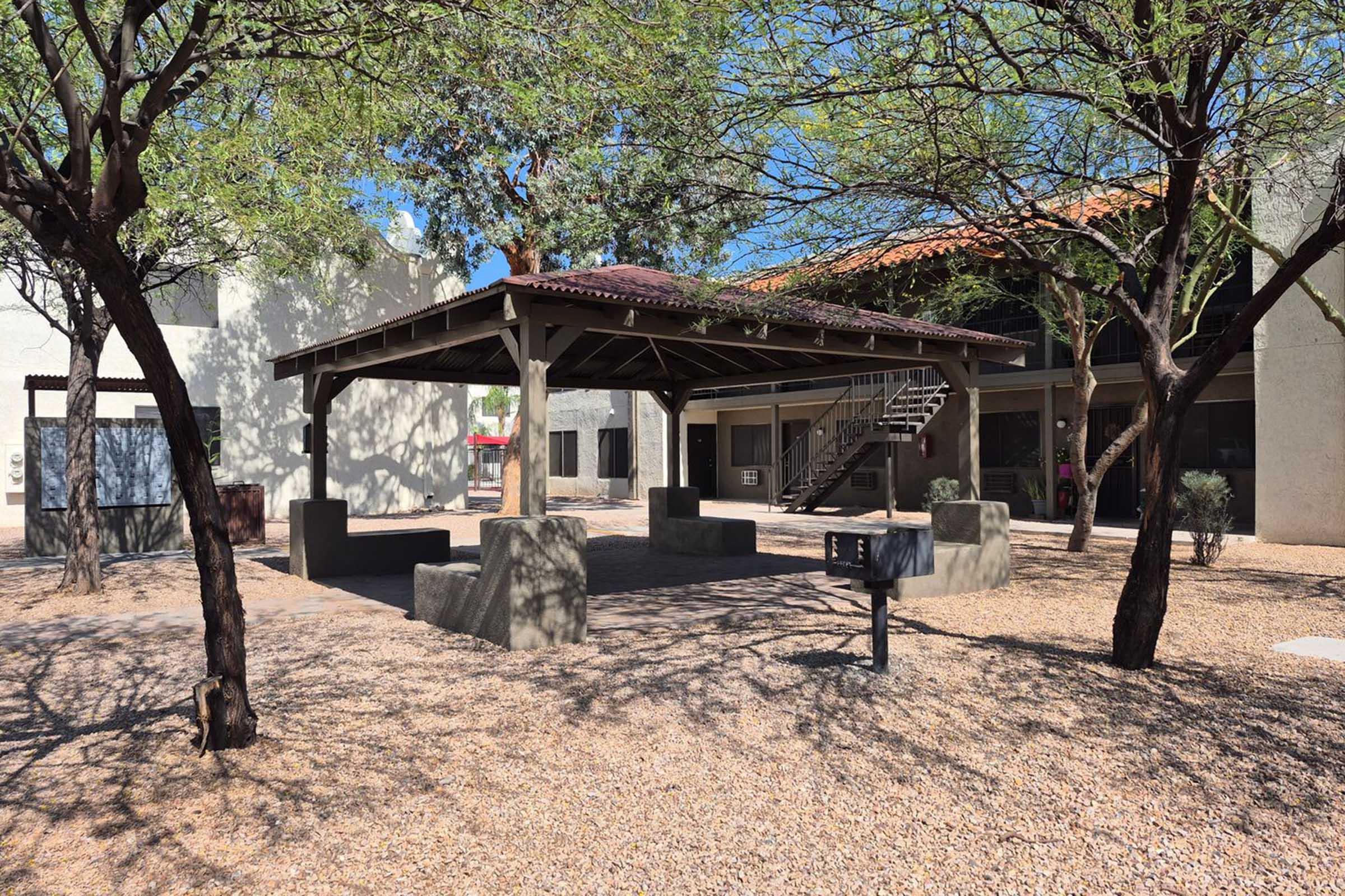 An outdoor courtyard featuring a gazebo-style structure with a red-tiled roof, surrounded by gravel and small trees. There are concrete seating areas and a barbecue grill, with nearby buildings visible in the background under a clear blue sky.