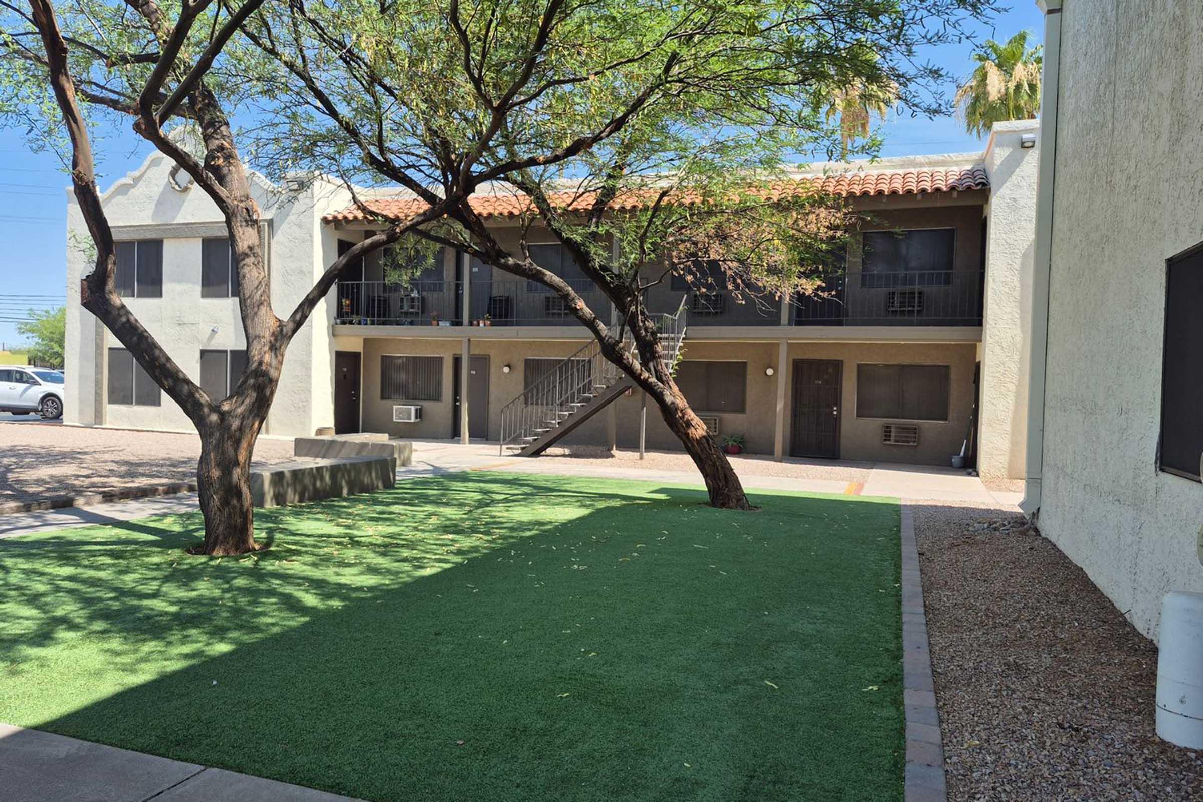 A view of a small courtyard featuring two trees and artificial grass, with a two-story apartment building in the background. The building has balconies and air conditioning units visible on the exterior. The scene is well-lit, suggesting a sunny day.