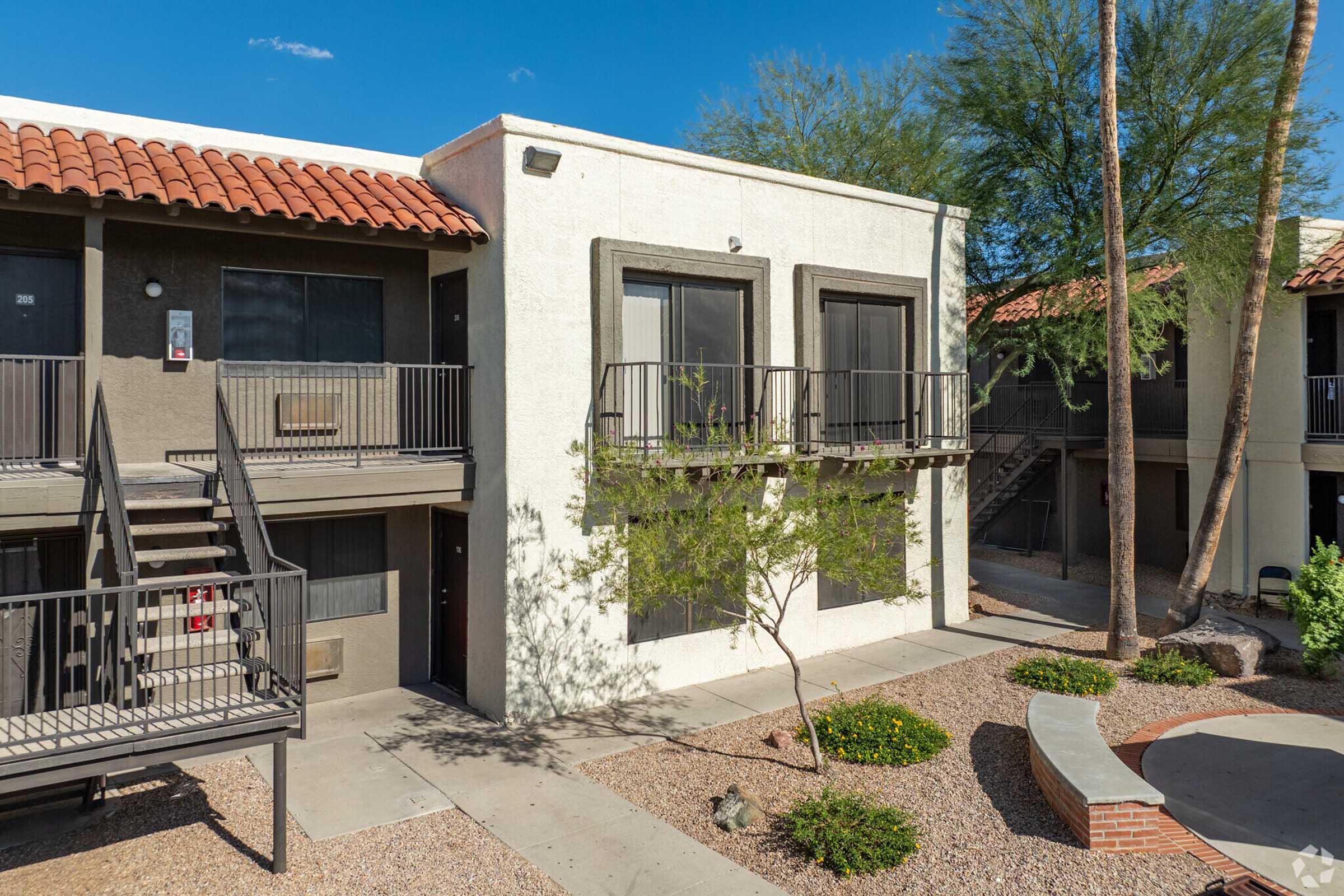 Two-story apartment complex with a stucco exterior and a red-tiled roof. The building features black metal balconies and a landscaped courtyard with gravel, small trees, and a circular stone seating area. Blue sky and palm trees are visible in the background.