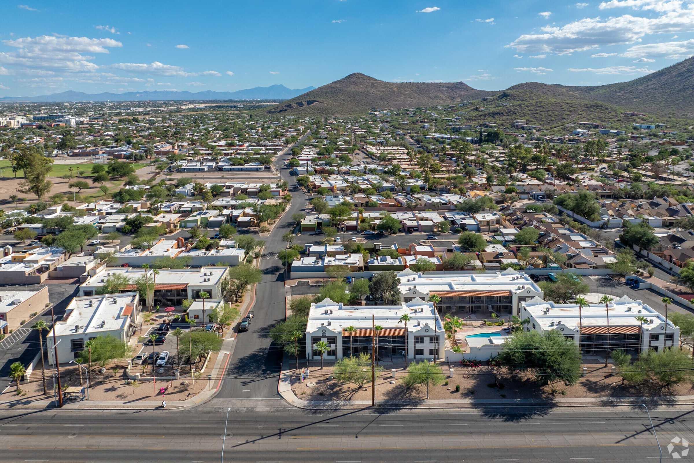 Aerial view of a suburban area featuring a mix of residential homes and low-rise buildings. Roads intersect, with vegetation and arid hills in the background. The landscape showcases a blend of urban development and natural greenery under a blue sky with scattered clouds.