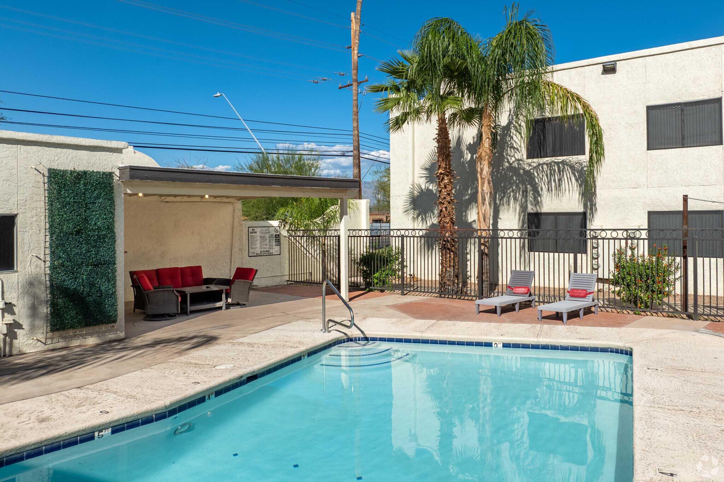 A serene swimming pool surrounded by palm trees and lounge chairs, with a shaded seating area featuring red furniture. A fence encloses the pool area, and a beige building is visible in the background against a clear blue sky.
