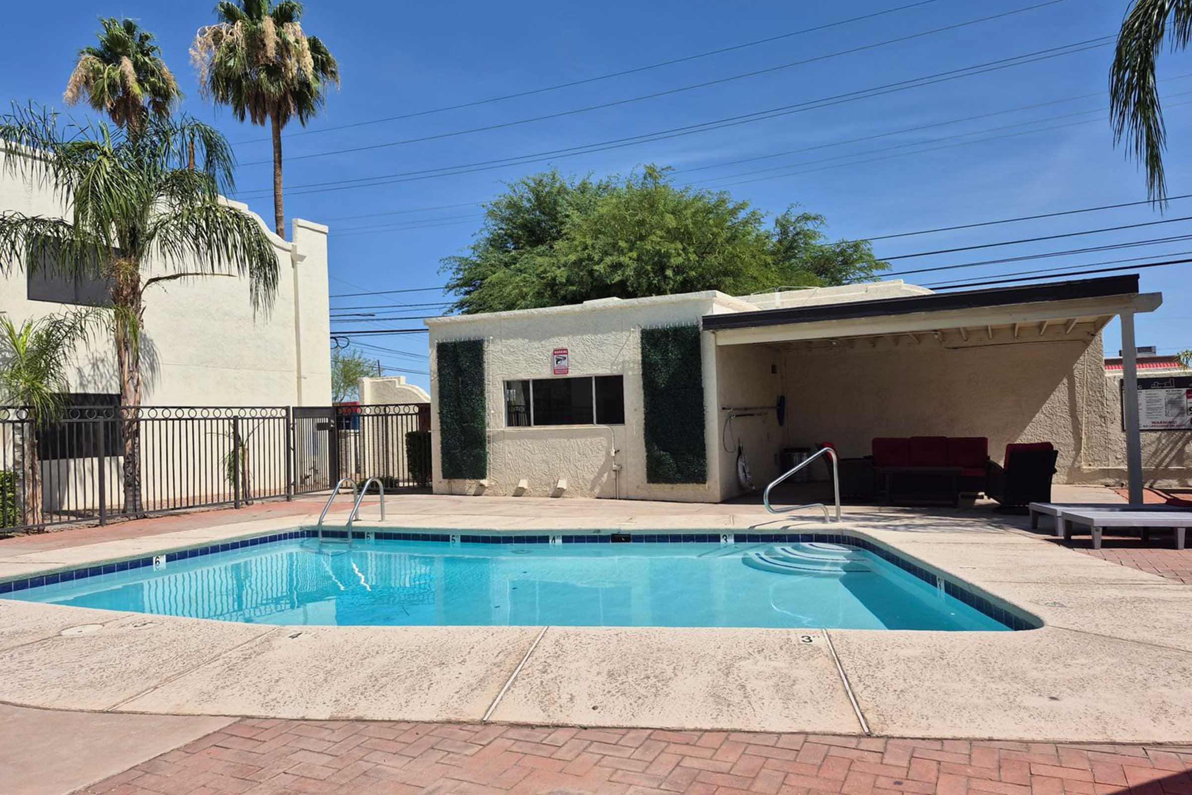 A backyard swimming pool with clear blue water, surrounded by a warm concrete deck. There are lounge chairs nearby and a shaded area with a seating arrangement under a small roof. In the background, palm trees are visible against a clear blue sky.